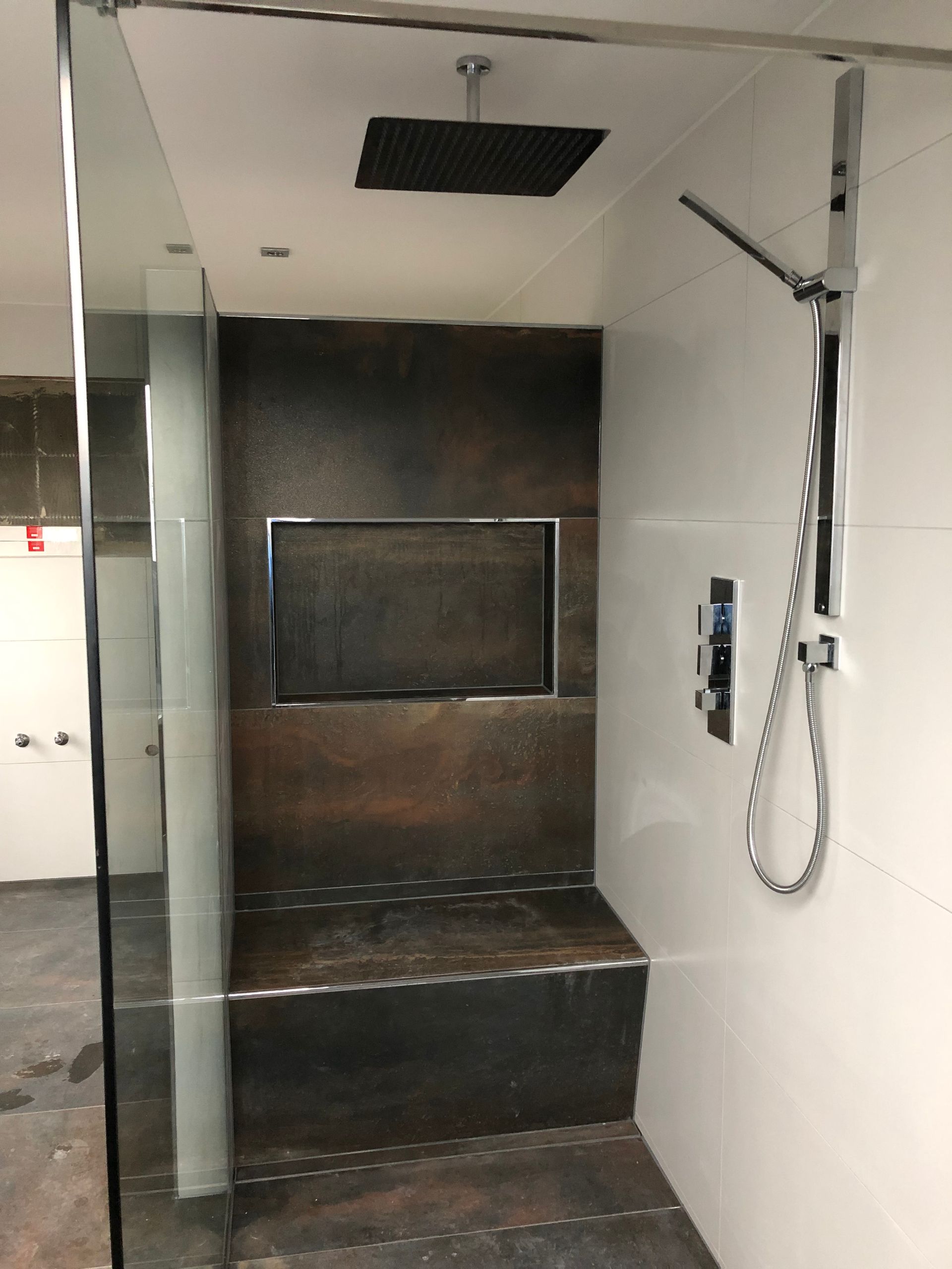Moderne begehbare Dusche mit dunkelfarbiger Sitzbank, Nische und Akzentwand. Weiß geflieste Wände und eine Glastür sind vorhanden.