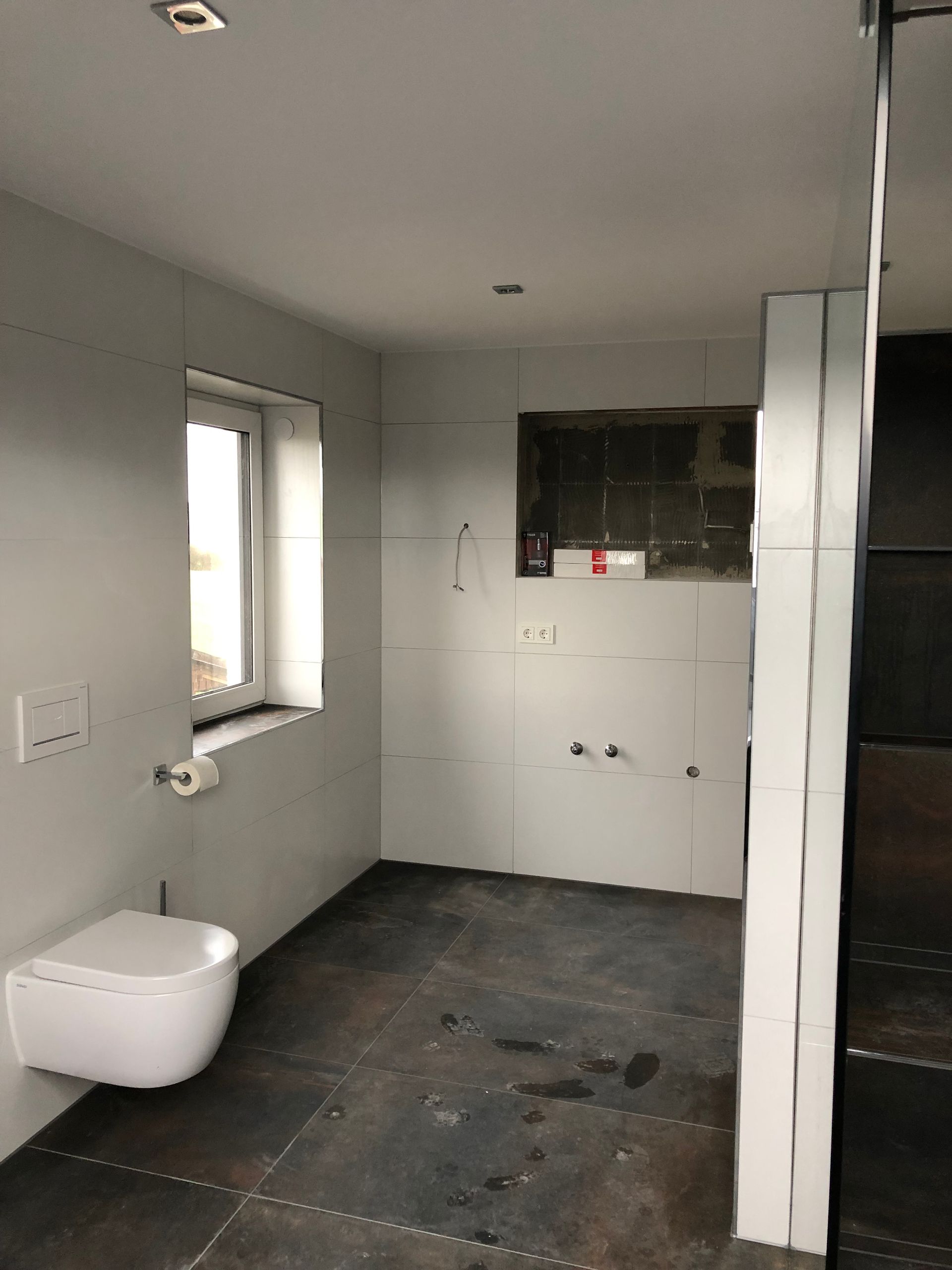 Badezimmer in Renovierung mit wandhängendem WC, Fenster und dunklem Boden.