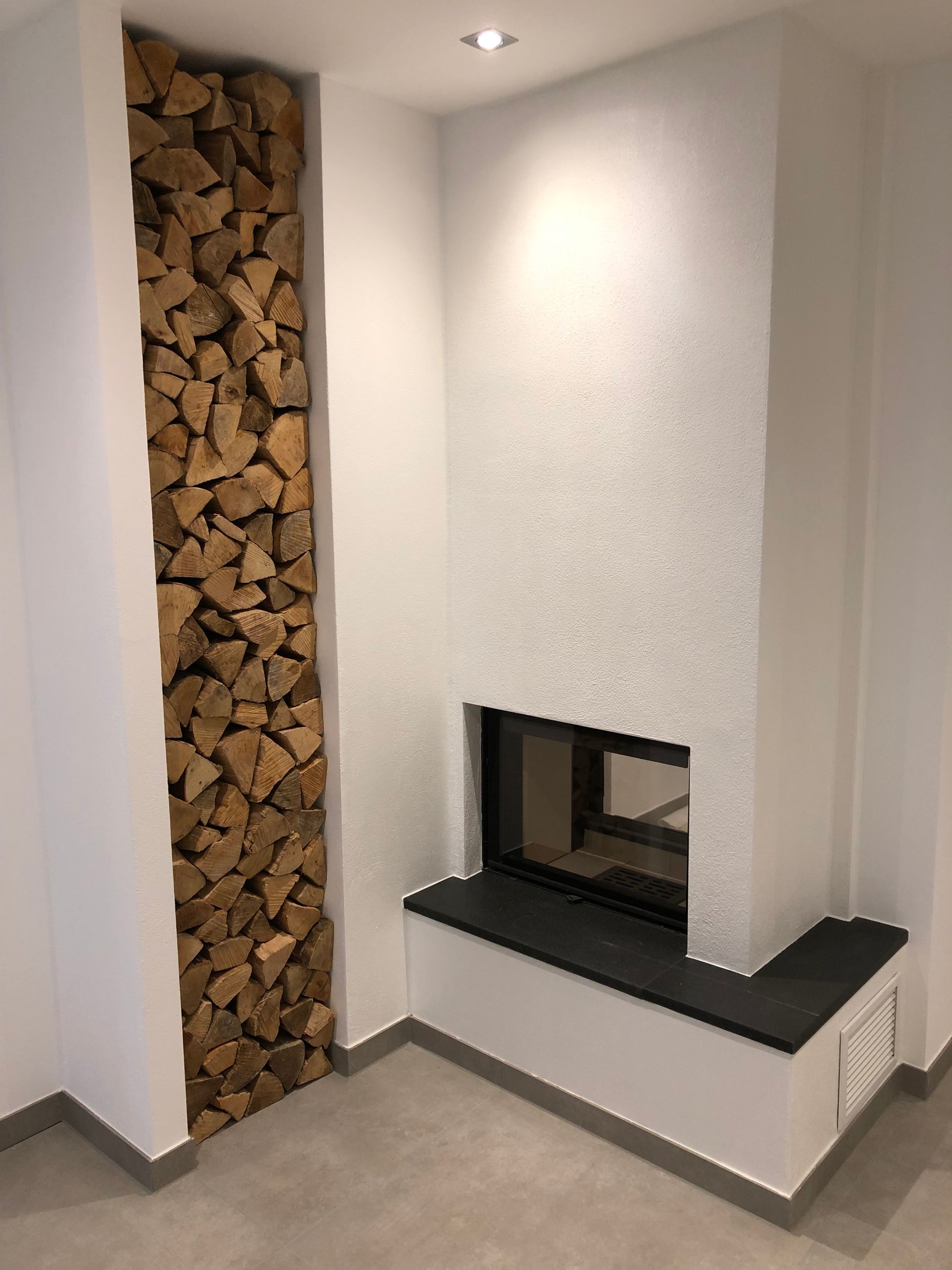 Kamin mit Holzlager auf der linken Seite und weiß strukturierter Wand.