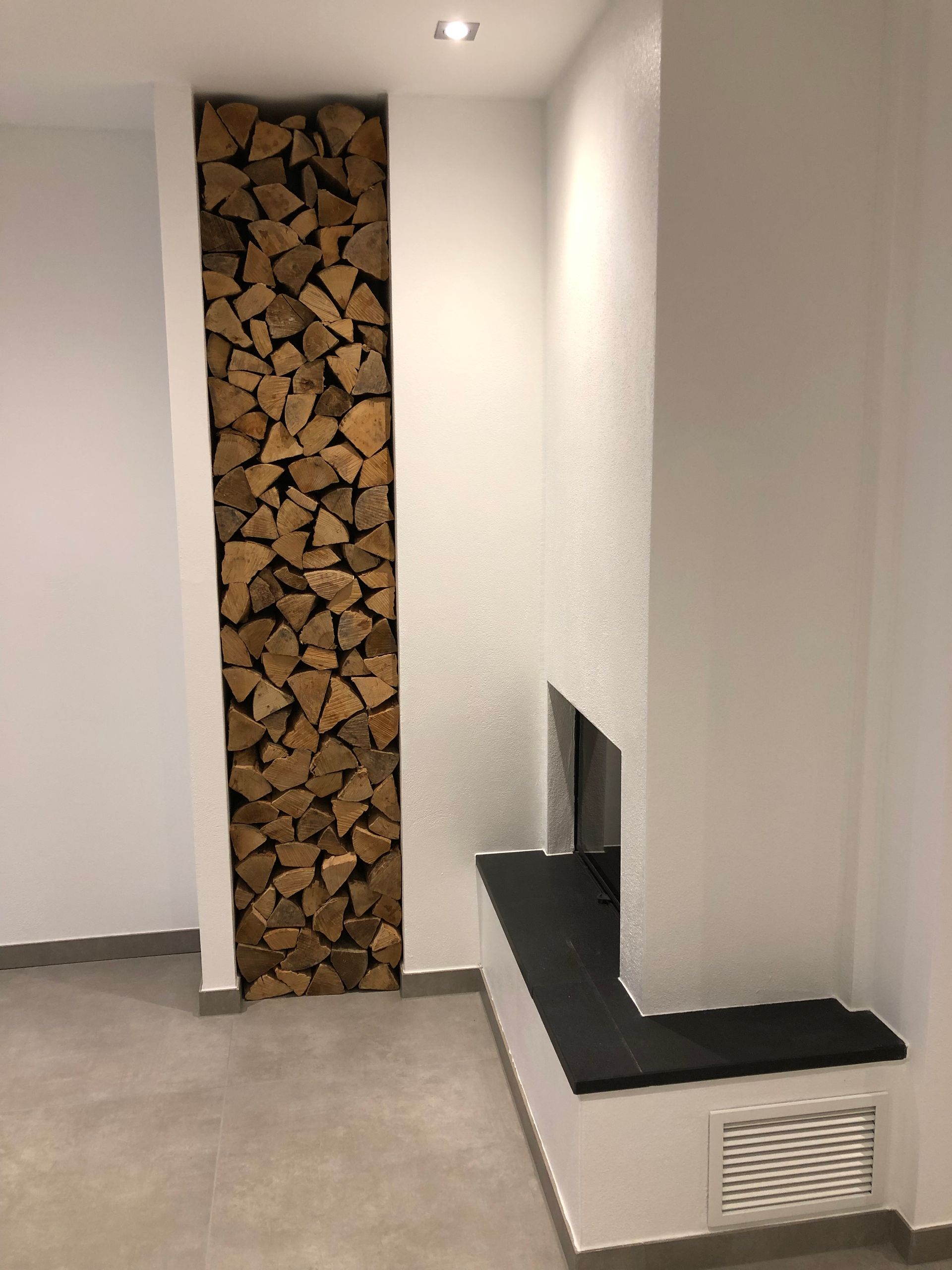 Brennholznische neben einem Kamin mit dunkler Feuerstelle; weiße Wände, grauer Fliesenboden.