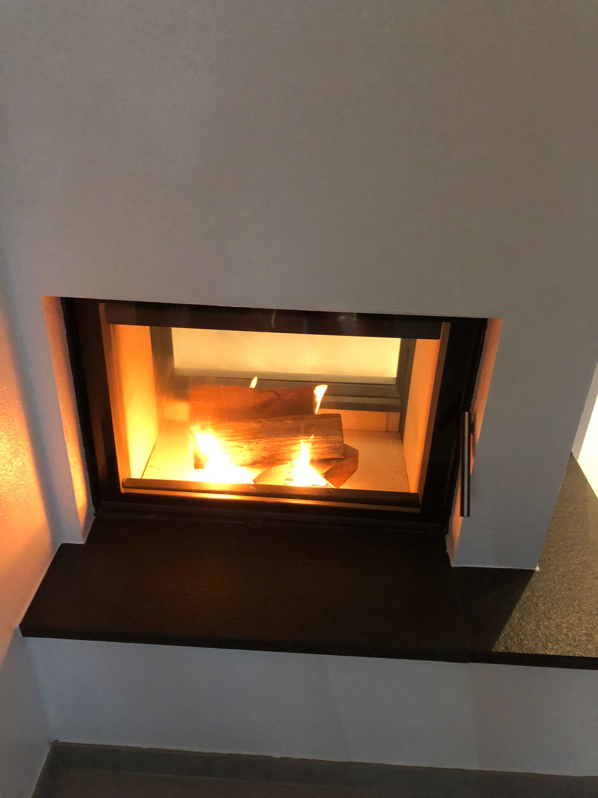 Kamin mit brennenden Holzscheiten im Inneren, in eine weiße Wand eingelassen, darunter ein dunkler Sims.