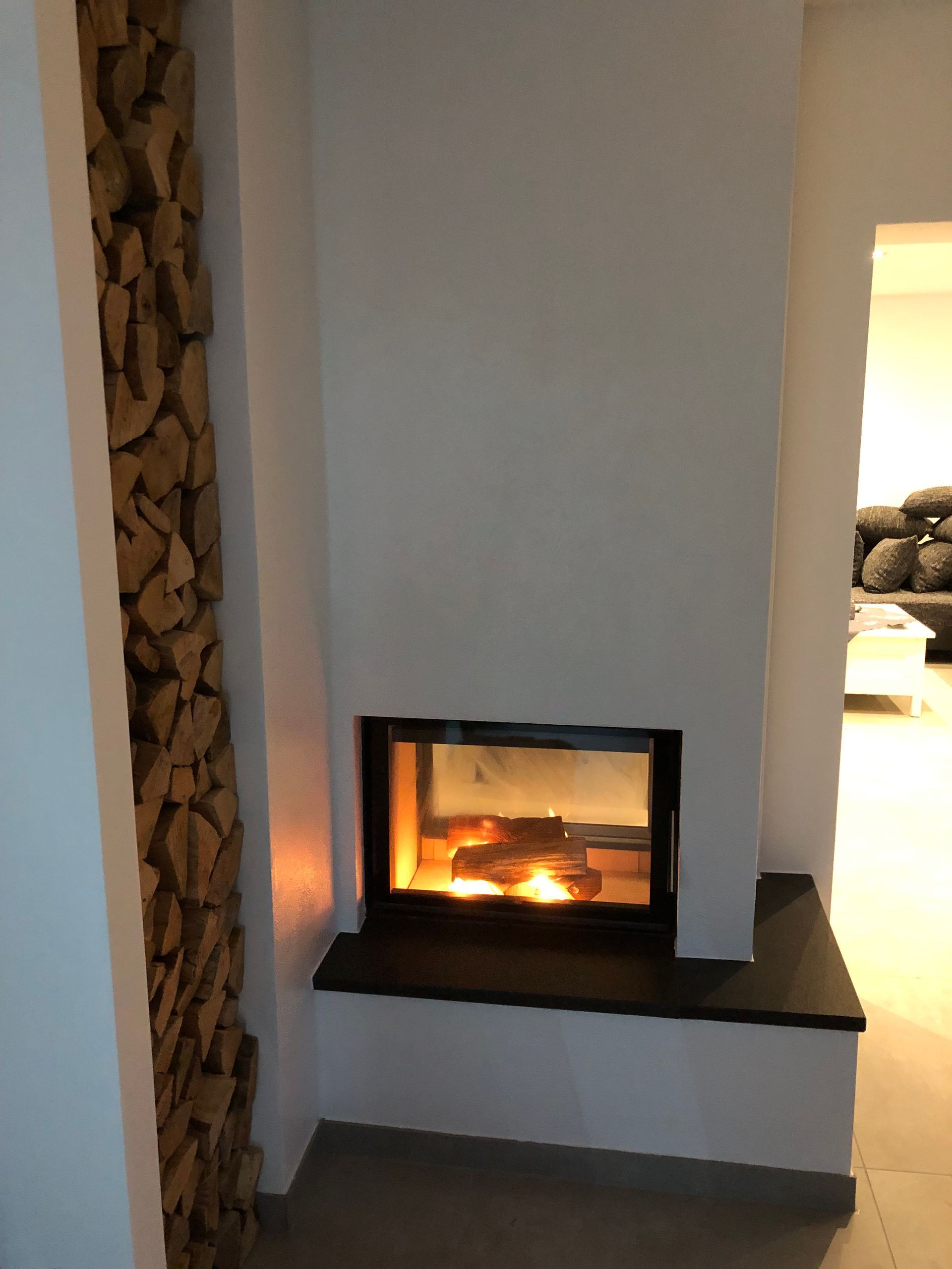 Kamin mit brennenden Holzscheiten, flankiert von gestapeltem Brennholz und weißen Wänden.