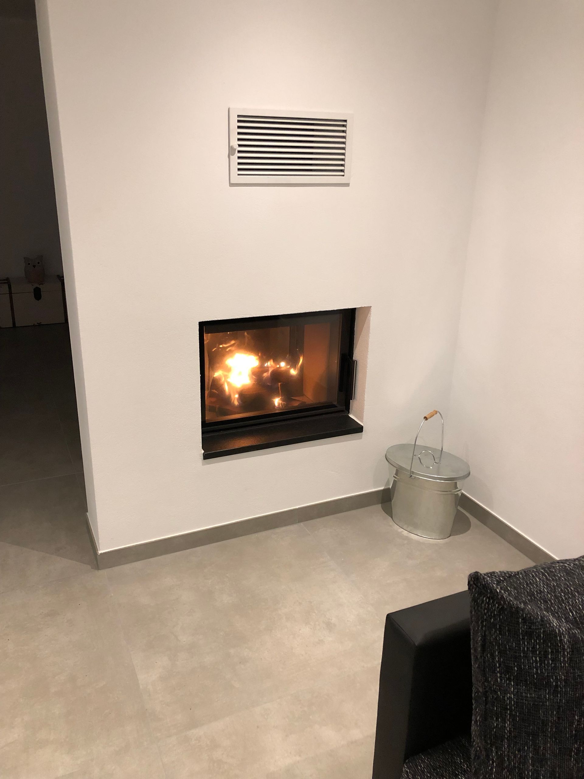 Kamin in einer weißen Wandnische, brennendes Feuer, Metalleimer daneben, darüber Abzugsöffnung.