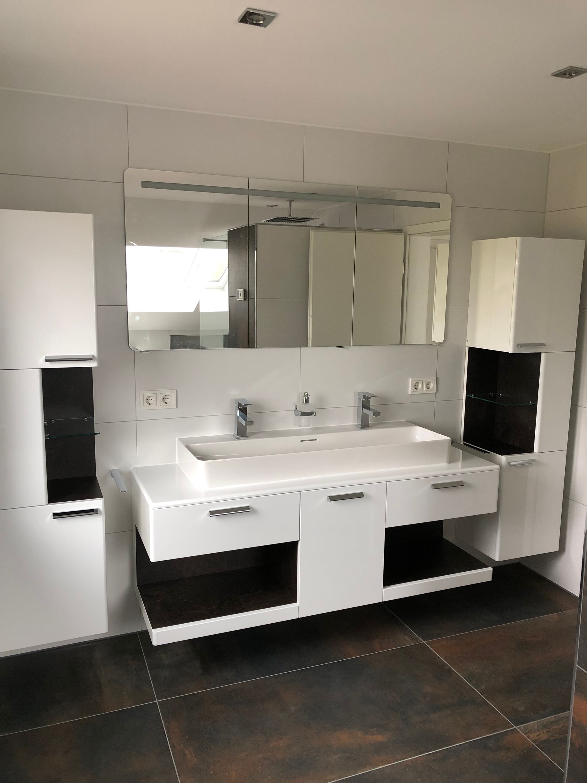 Modernes weißes Badezimmer mit Doppelwaschbecken, Spiegel und Aufbewahrungsschränken auf dunklem Fliesenboden.