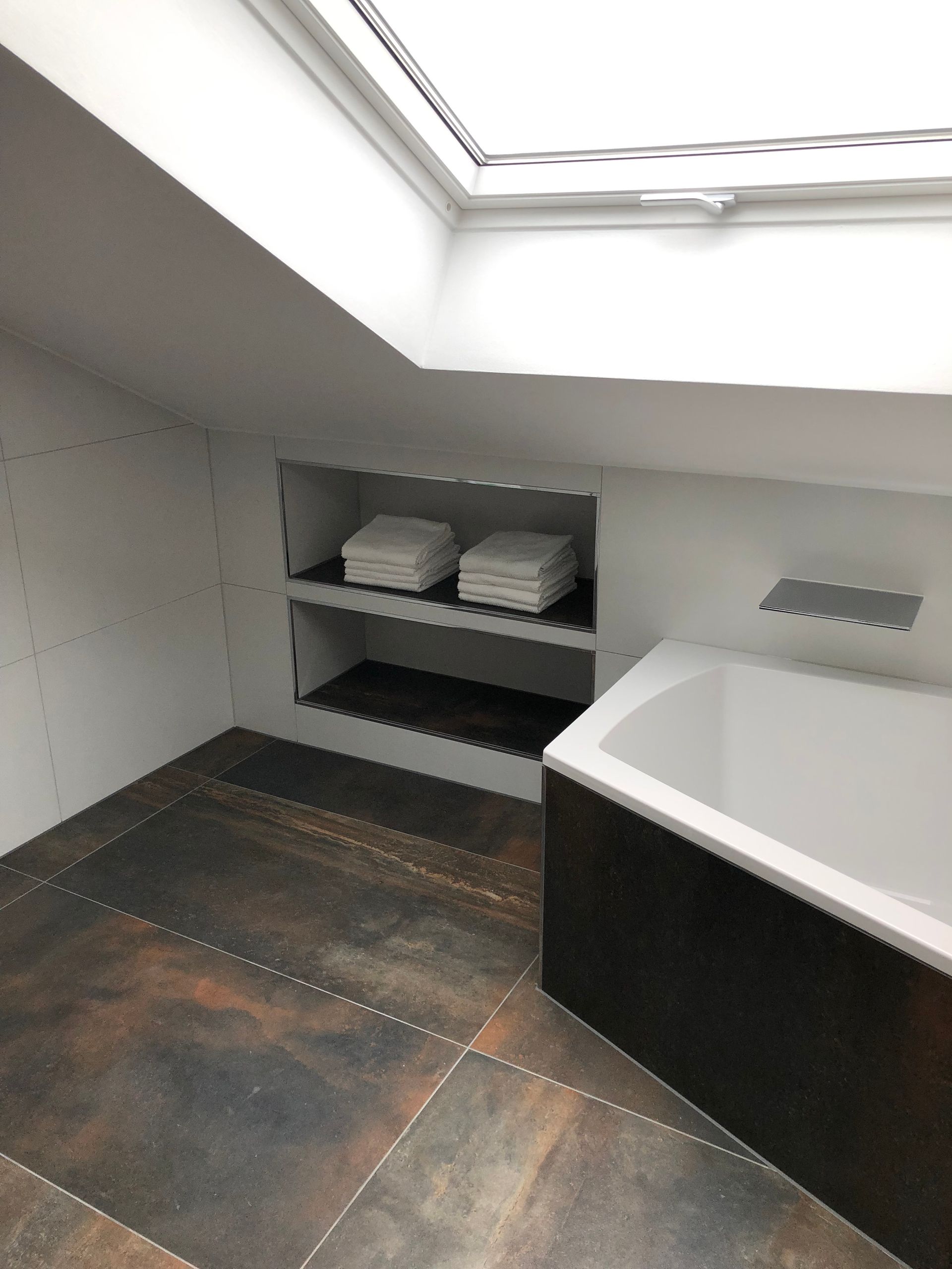 Badezimmer mit schräger Decke, Oberlicht, eingebauten Regalen mit Handtüchern, Badewanne und strukturiertem dunklem Boden.