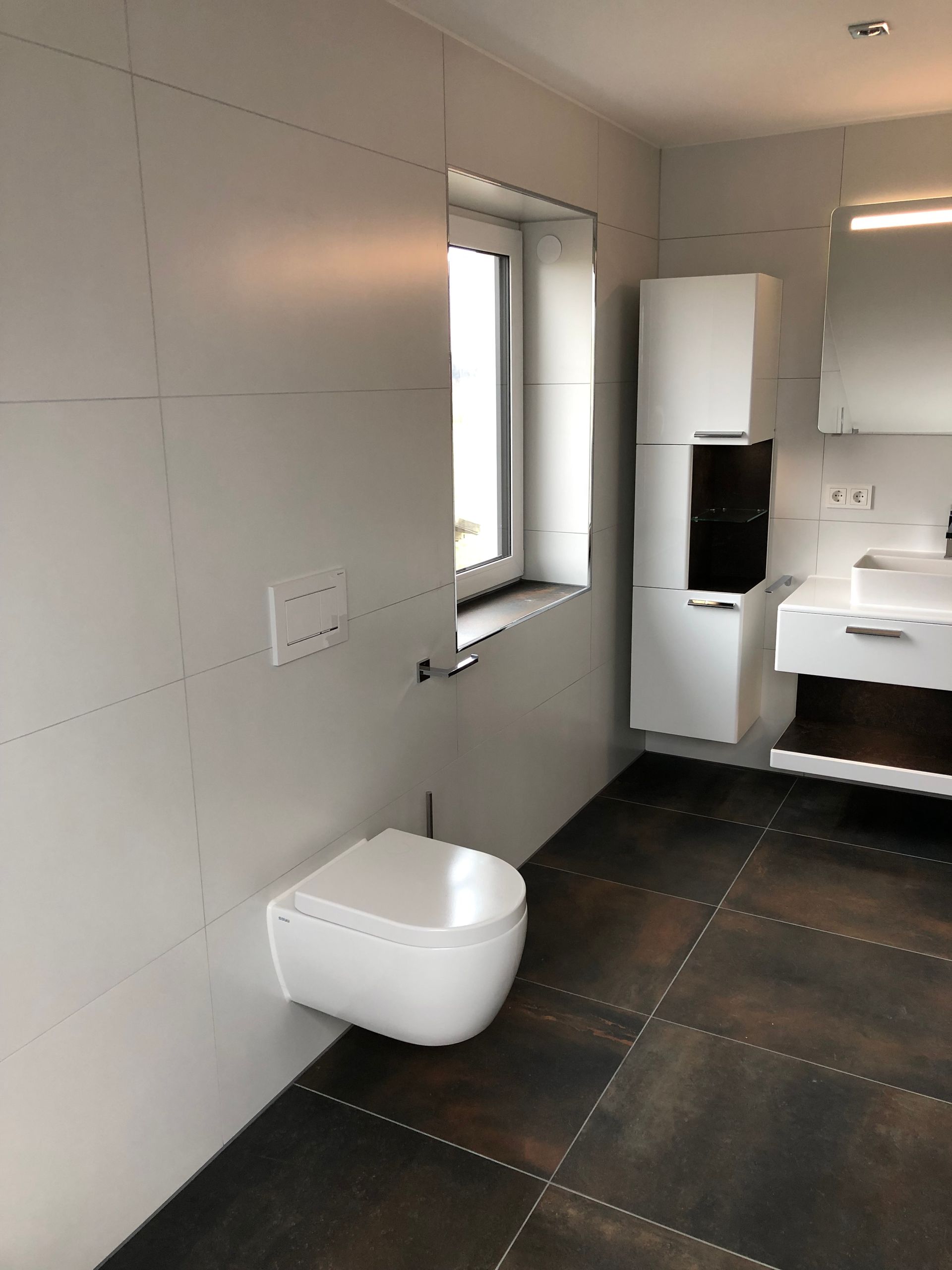 Modernes Badezimmer mit weißen Wänden, dunklem Fliesenboden und weißer Toilette unter einem Fenster.