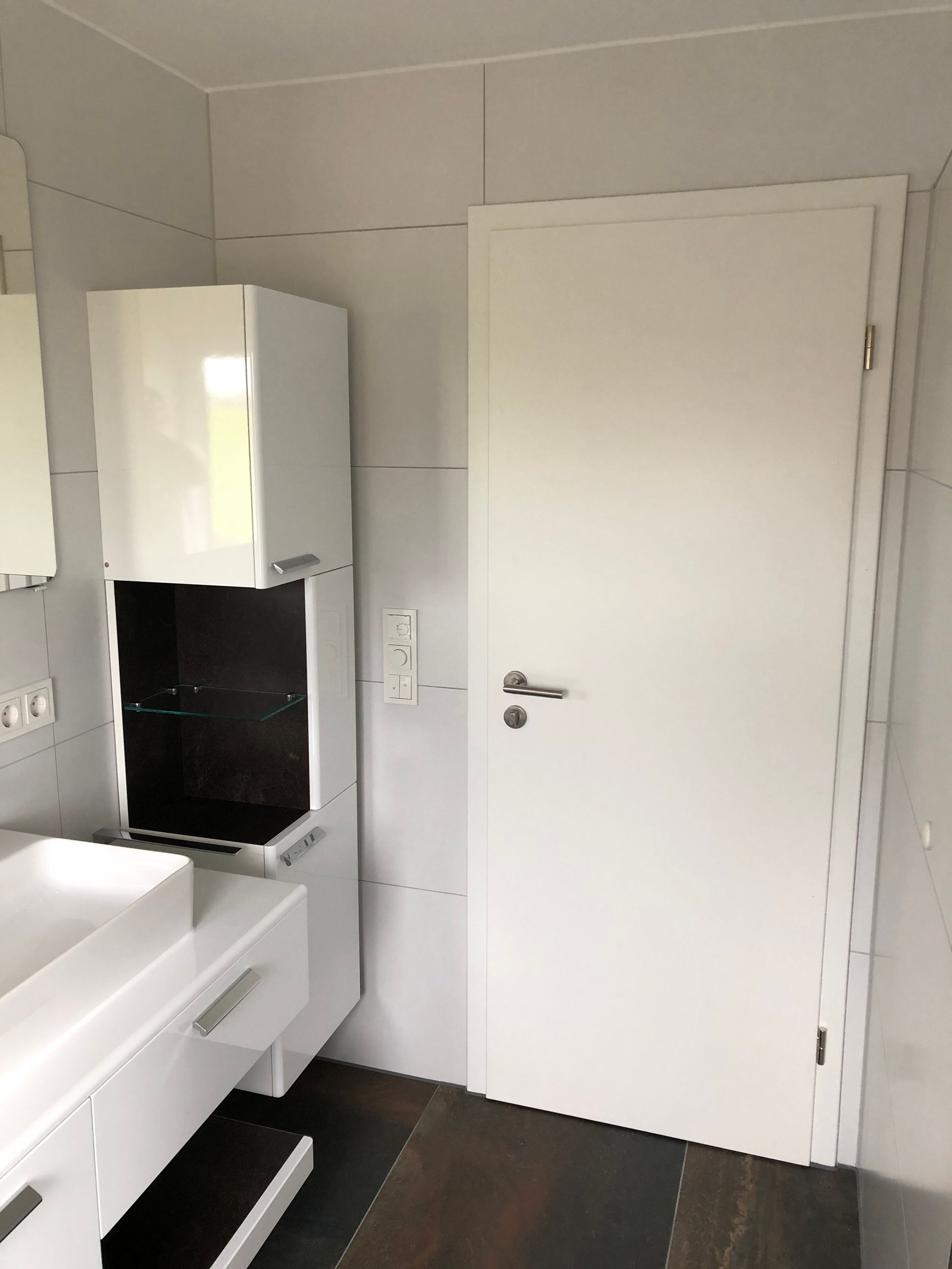 Badezimmer mit weißen Schränken, Waschbecken und geschlossener Tür. Dunkler Holzboden, hellgrau geflieste Wände.