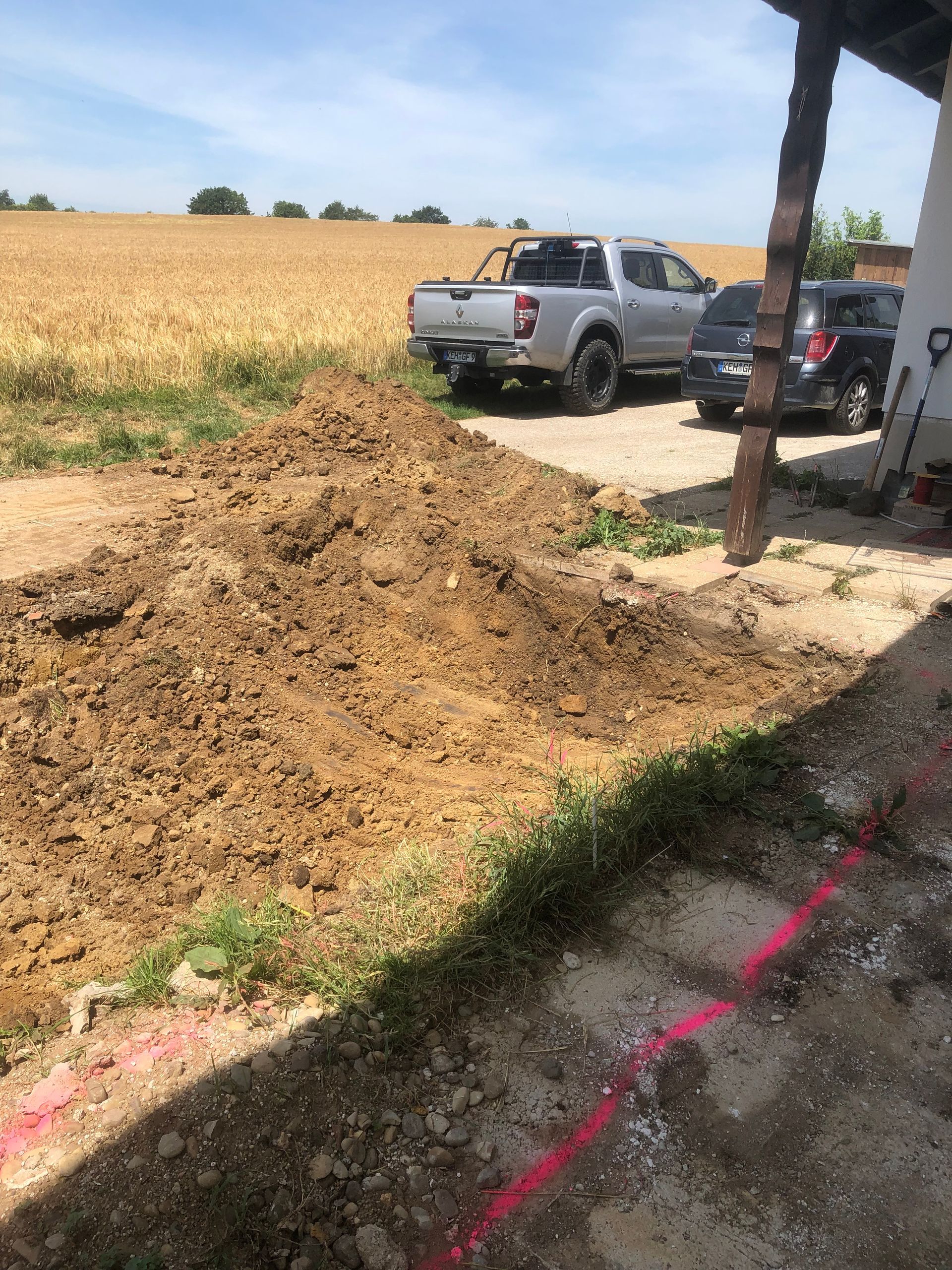 Erdhaufen neben einem Gebäude mit rosa Linien, einem LKW und einem Feld im Hintergrund. Sonniger Tag.