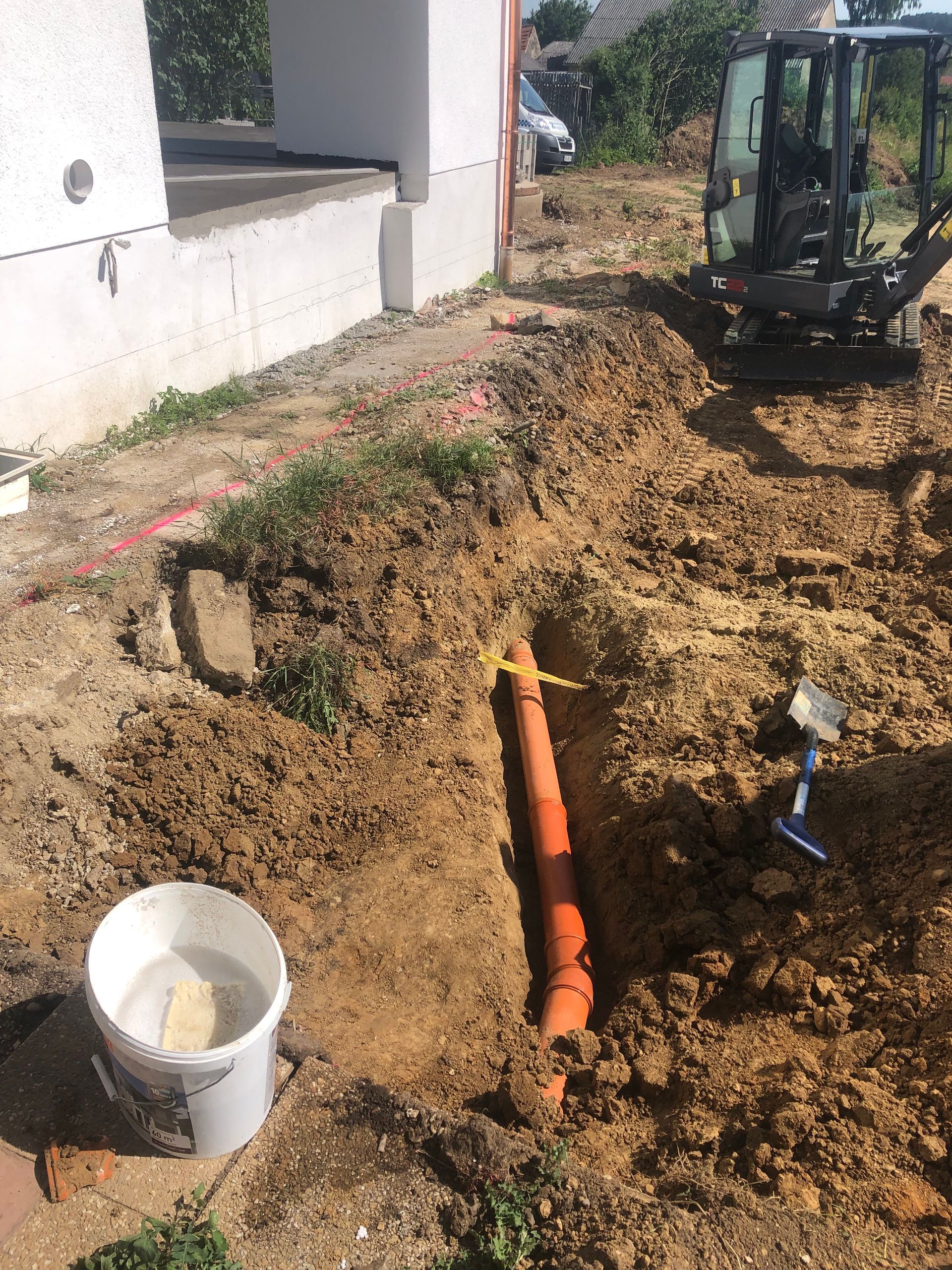 Graben in der Nähe eines Gebäudes mit orangefarbenem Rohr und kleinem Bagger im Hintergrund. Weißer Eimer und Schaufel stehen in der Nähe.