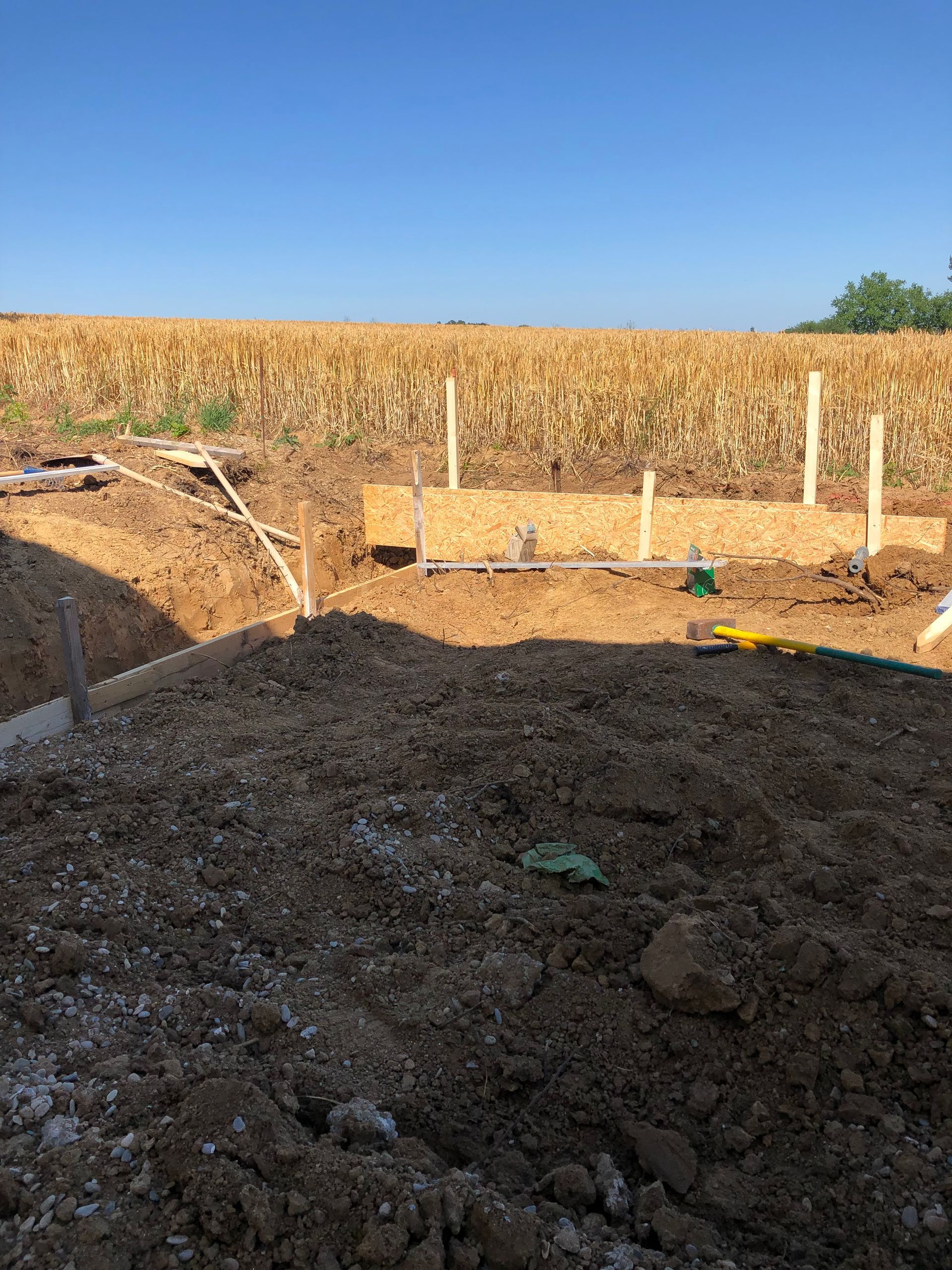 Baustelle: Erdhaufen, Holzpfosten, ein Graben und ein Feld mit Getreide unter blauem Himmel.
