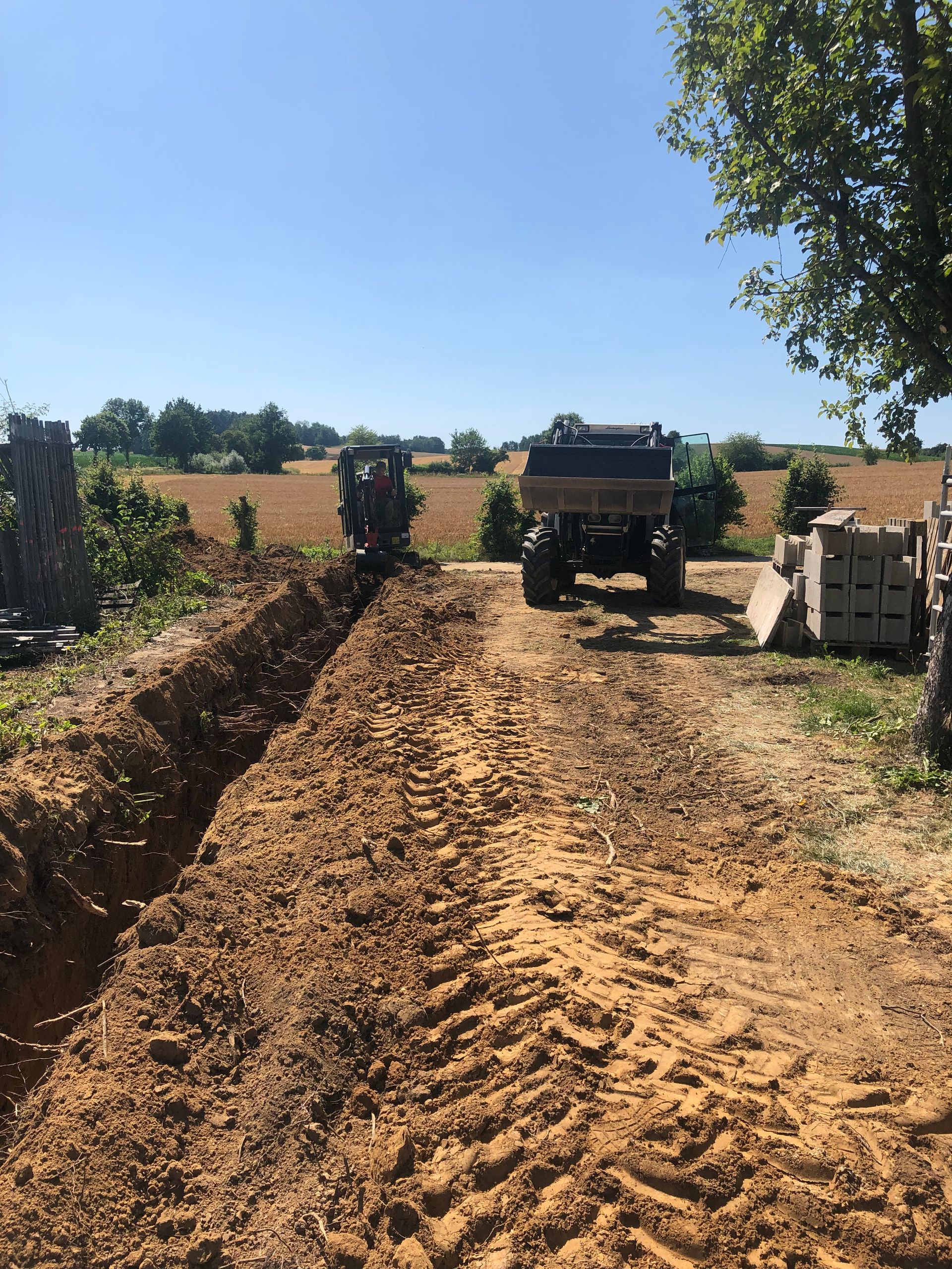 Ein für Bauarbeiten ausgehobener Graben mit zwei Fahrzeugen in der Nähe unter blauem Himmel und Bäumen.