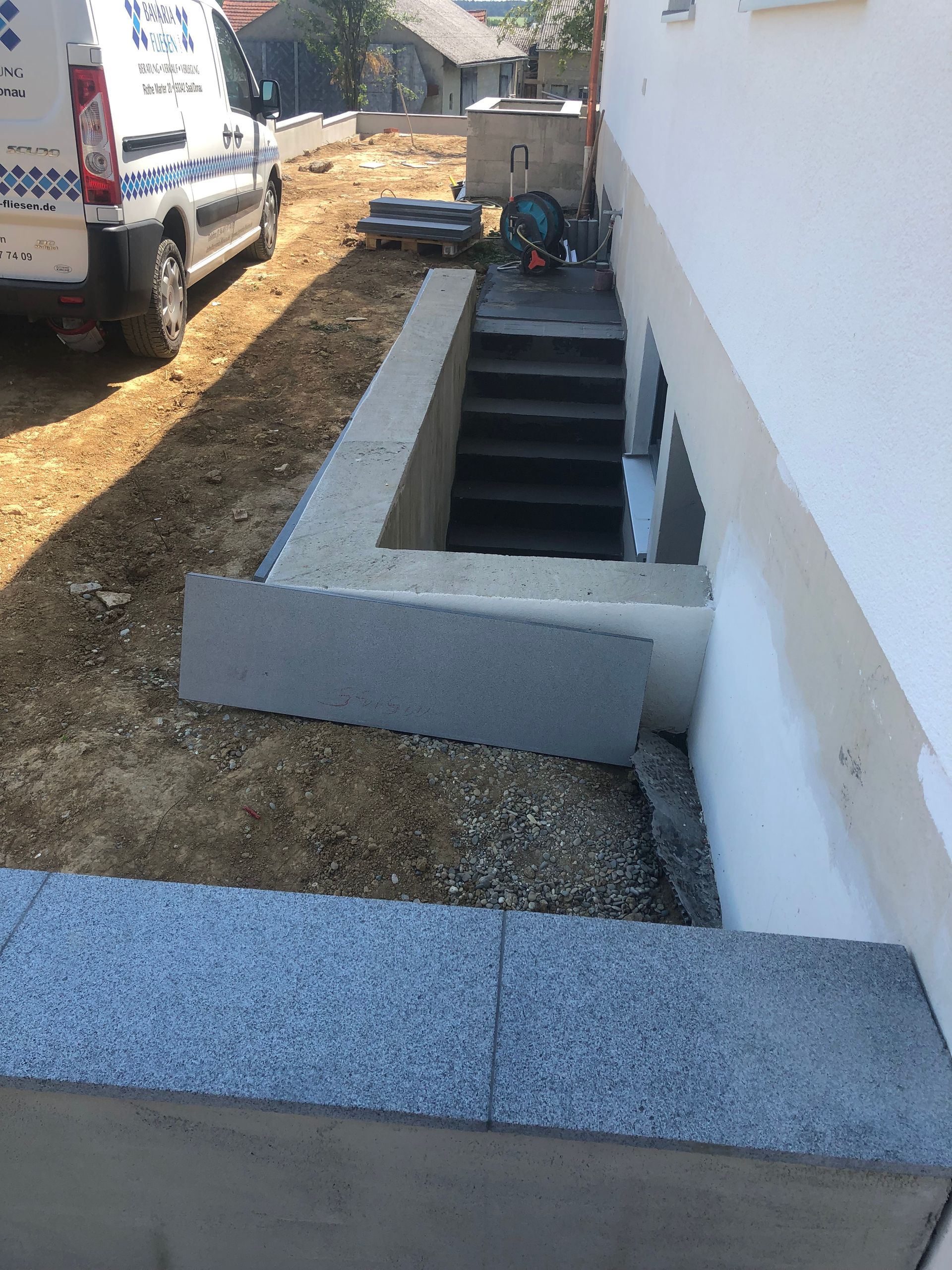 Eine Betontreppe führt hinunter zu einem Kellereingang; ein weißes Gebäude und eine Landschaftsgestaltung sind vorhanden.