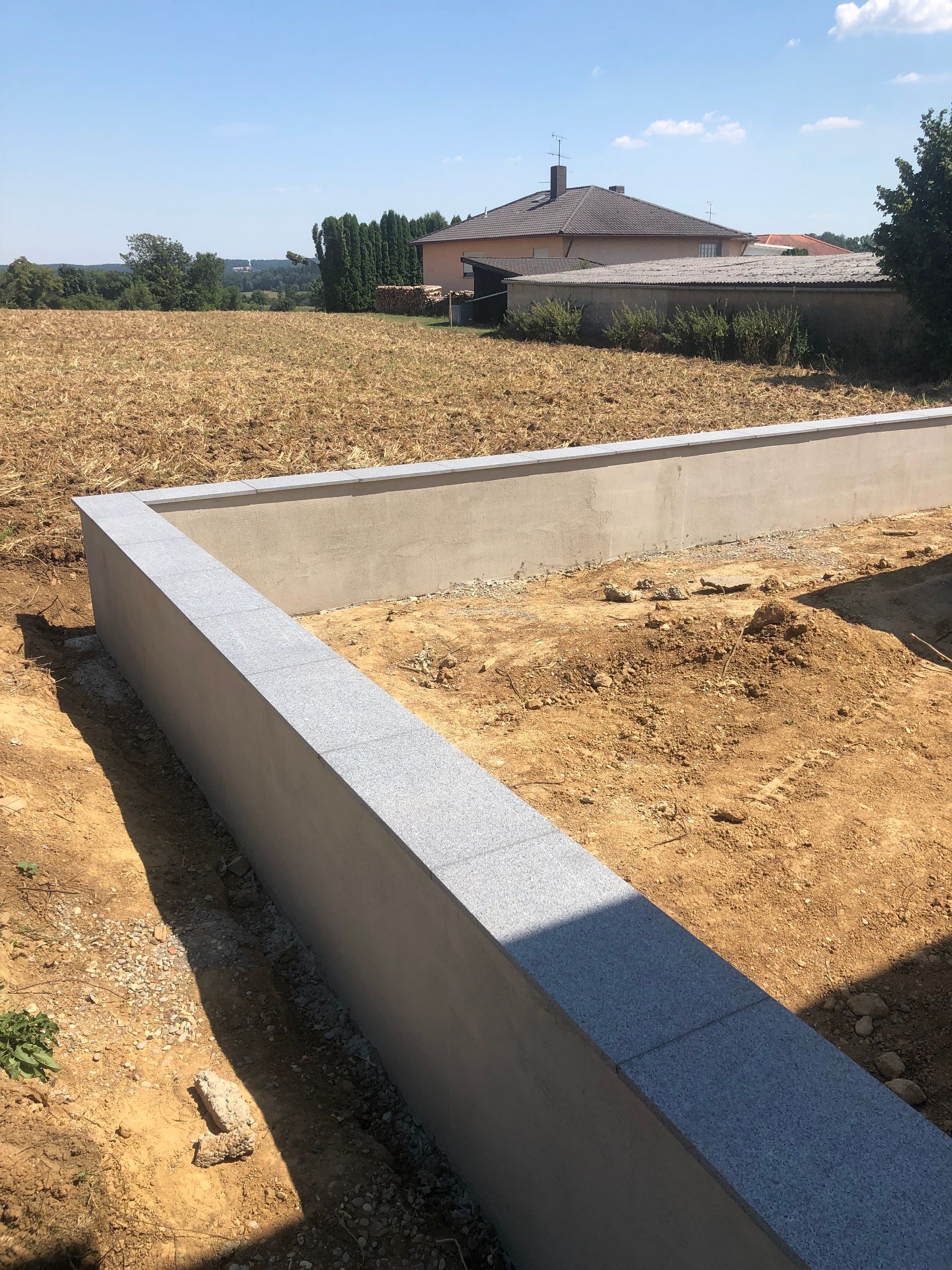 Eine neu errichtete graue Stützmauer auf einem sonnigen Feld mit einem Haus im Hintergrund.