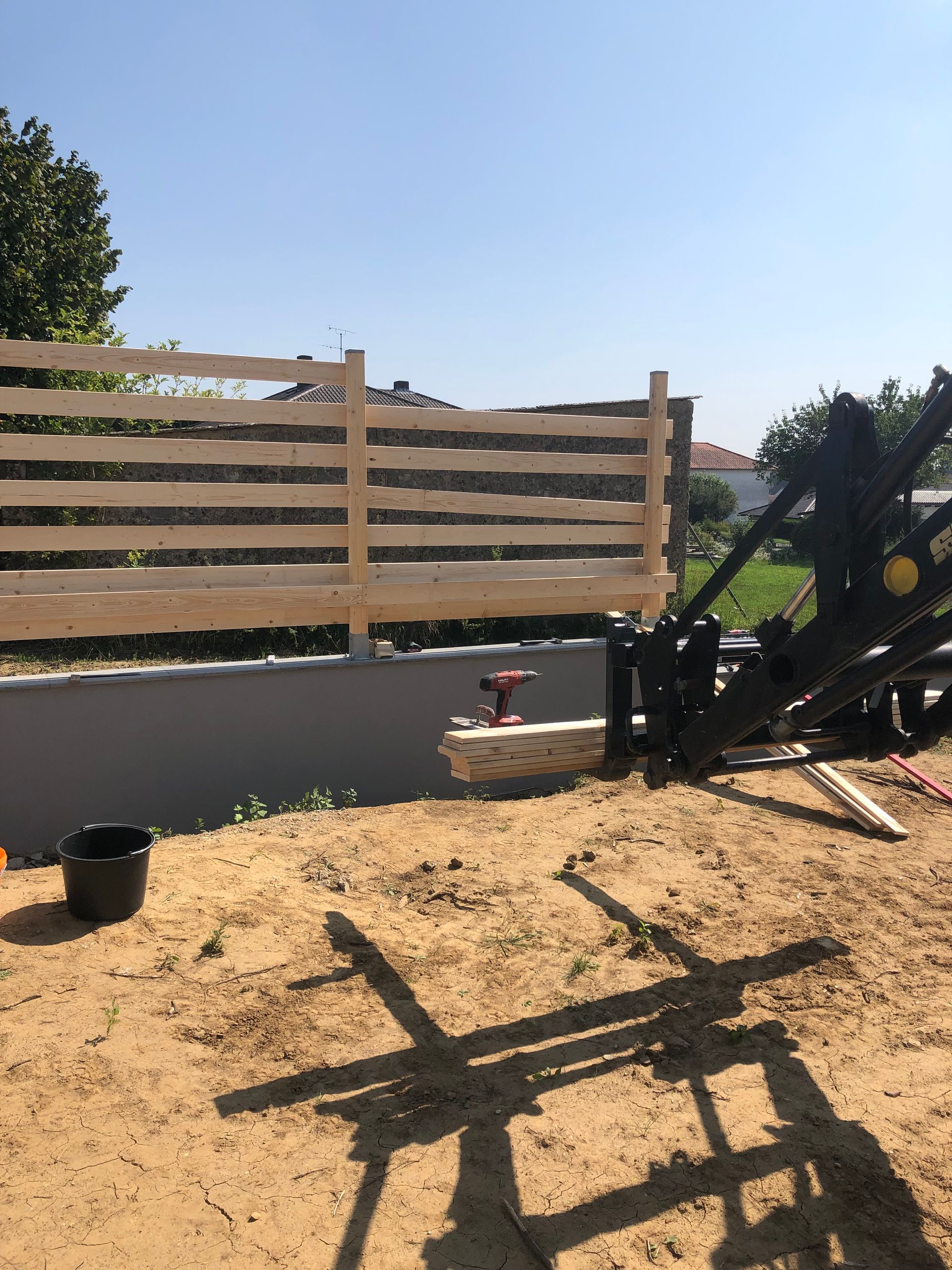 Zaunbau im Gange: Holzzaun, Sand, Werkzeuge, Kran, sonniger Tag.