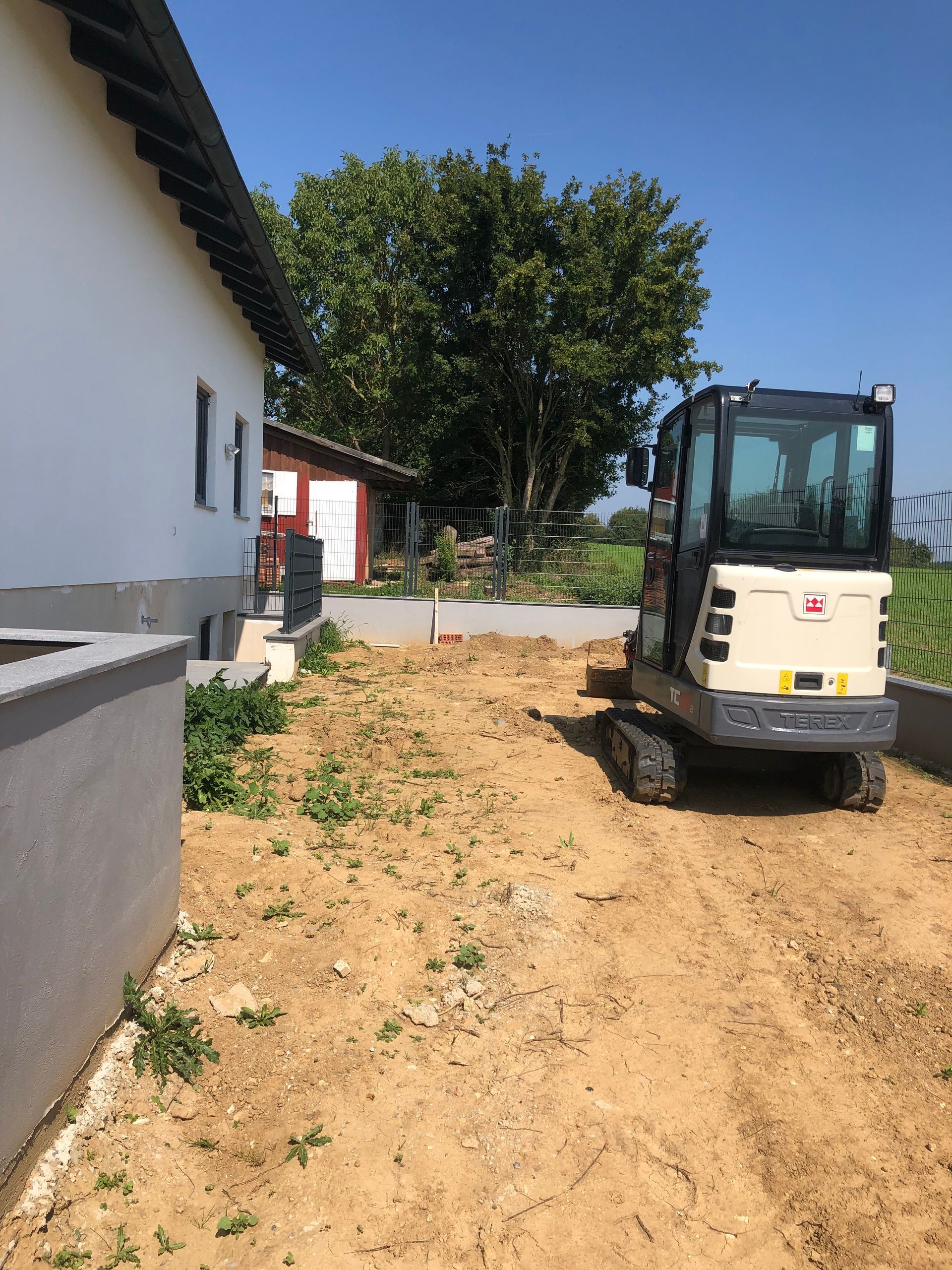 Minibagger auf einem Feldweg neben einem weißen Gebäude und einem kleinen roten Schuppen unter blauem Himmel.