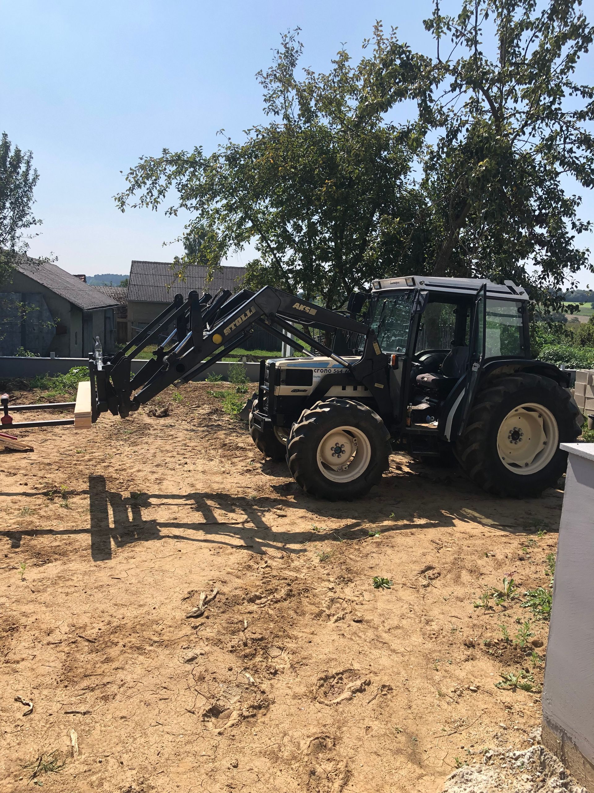 Traktor mit Frontlader auf einer Baustelle, der einen Holzbalken transportiert. Sonniger Tag.