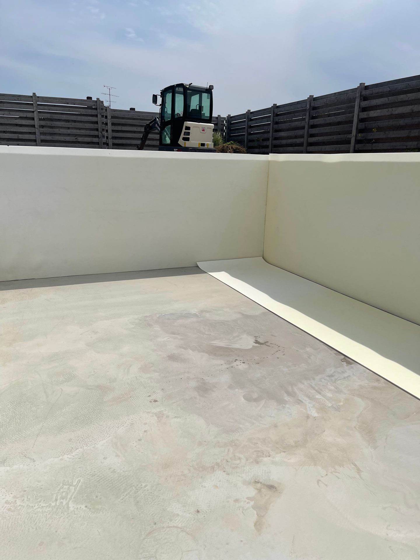 Ein kleines Baufahrzeug auf einer Betonfläche, in der Nähe von hellbraunen Wänden und einem Holzzaun.