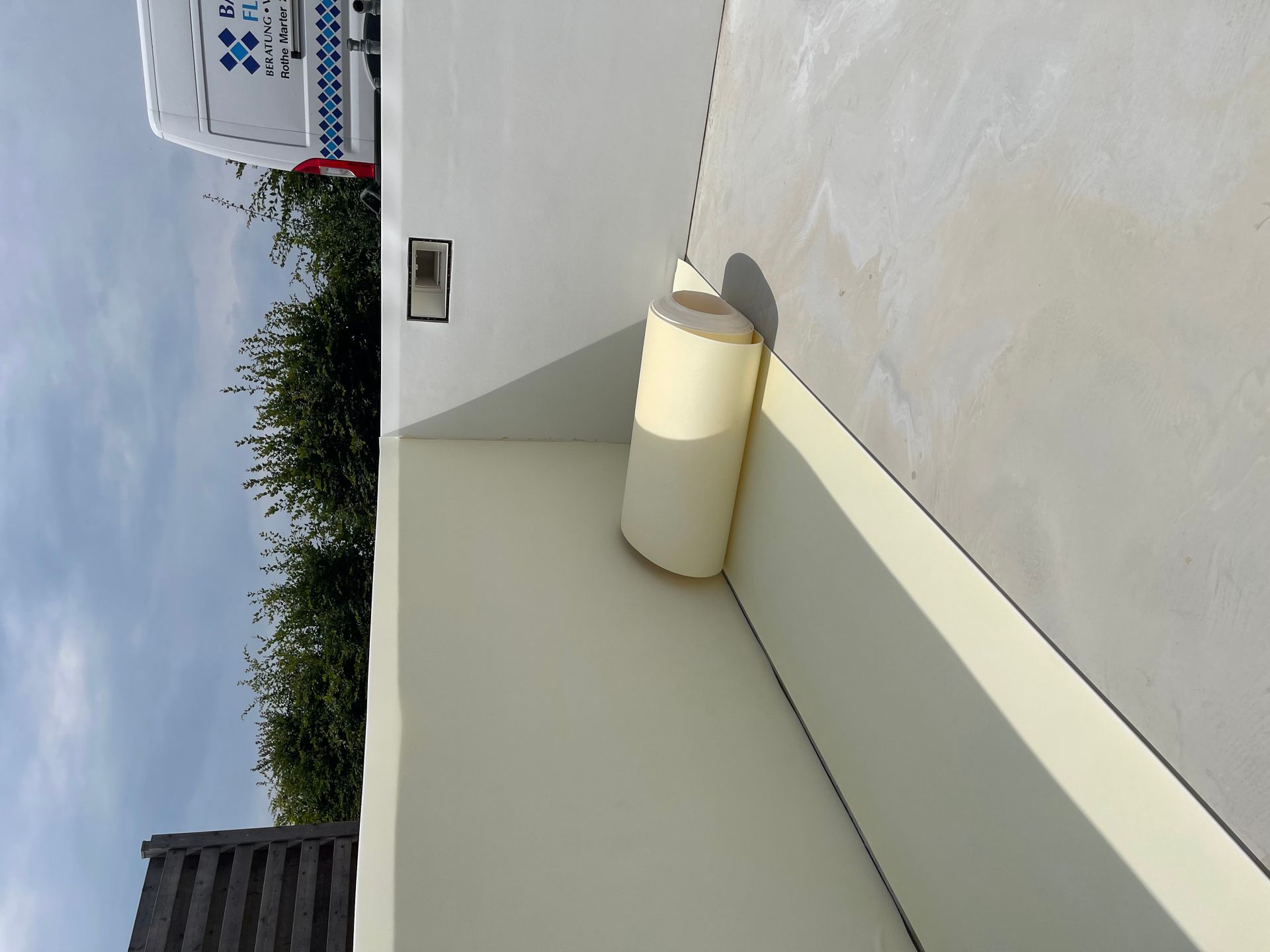 Eine Dämmstoffrolle lehnt an einer Wand neben einem Dach, im Hintergrund ist ein Lieferwagen zu sehen.