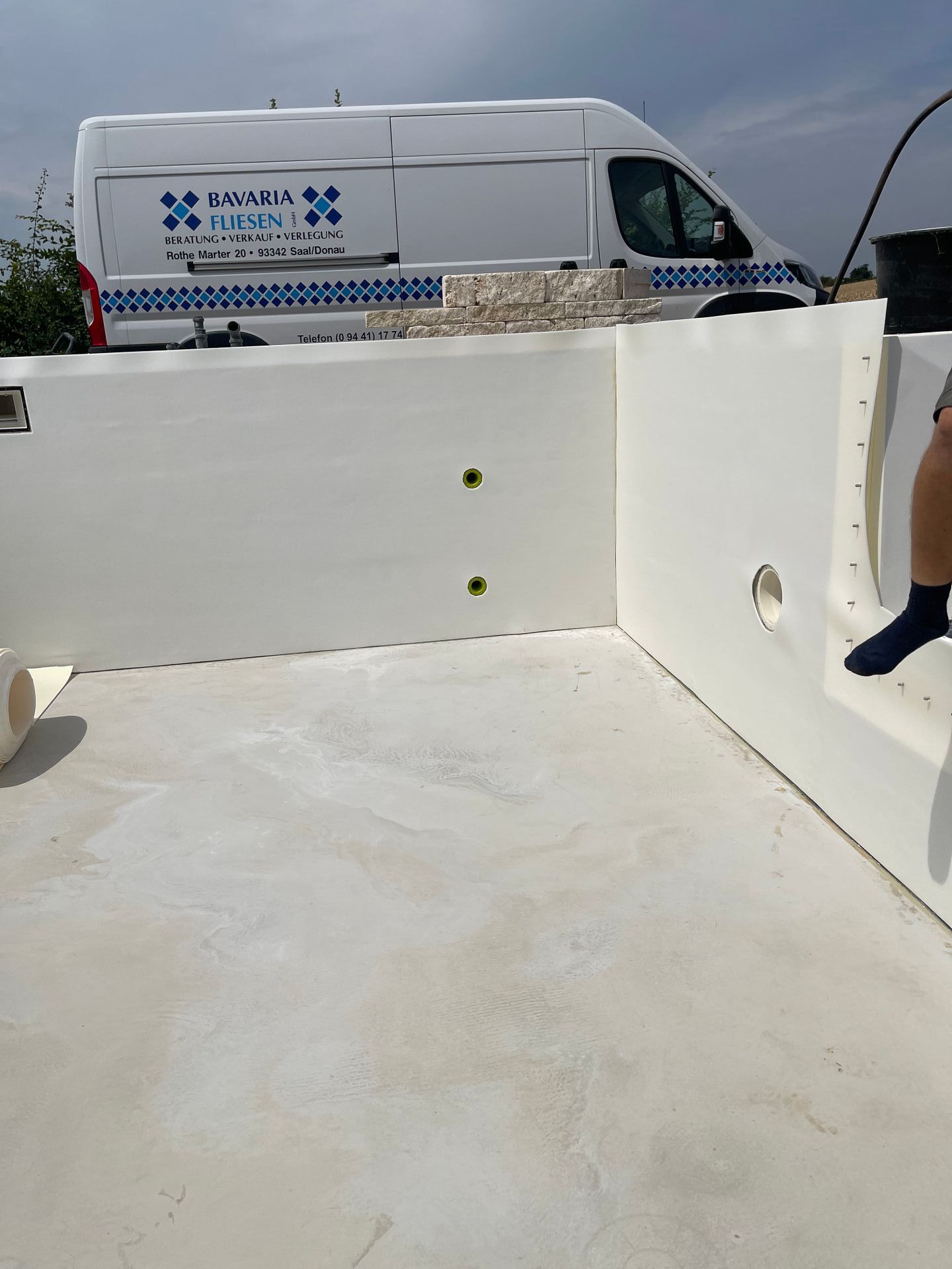 Weißer Pool im Bau, Lieferwagen mit Logo, Bein einer Person sichtbar, hellgraue Oberfläche.