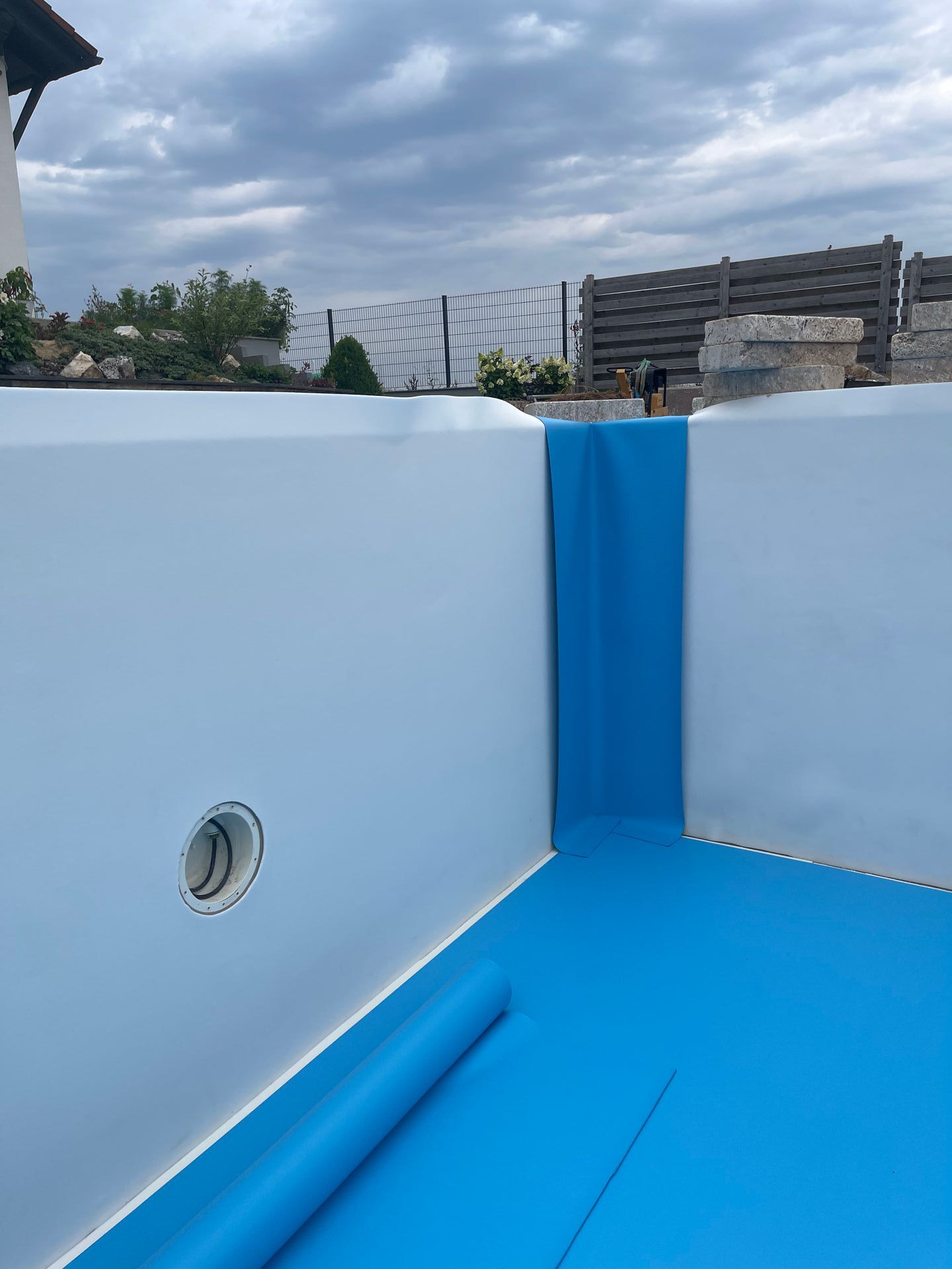 Innenansicht eines neuen, blau umrandeten Schwimmbeckens im Bau, darüber ein bewölkter Himmel.