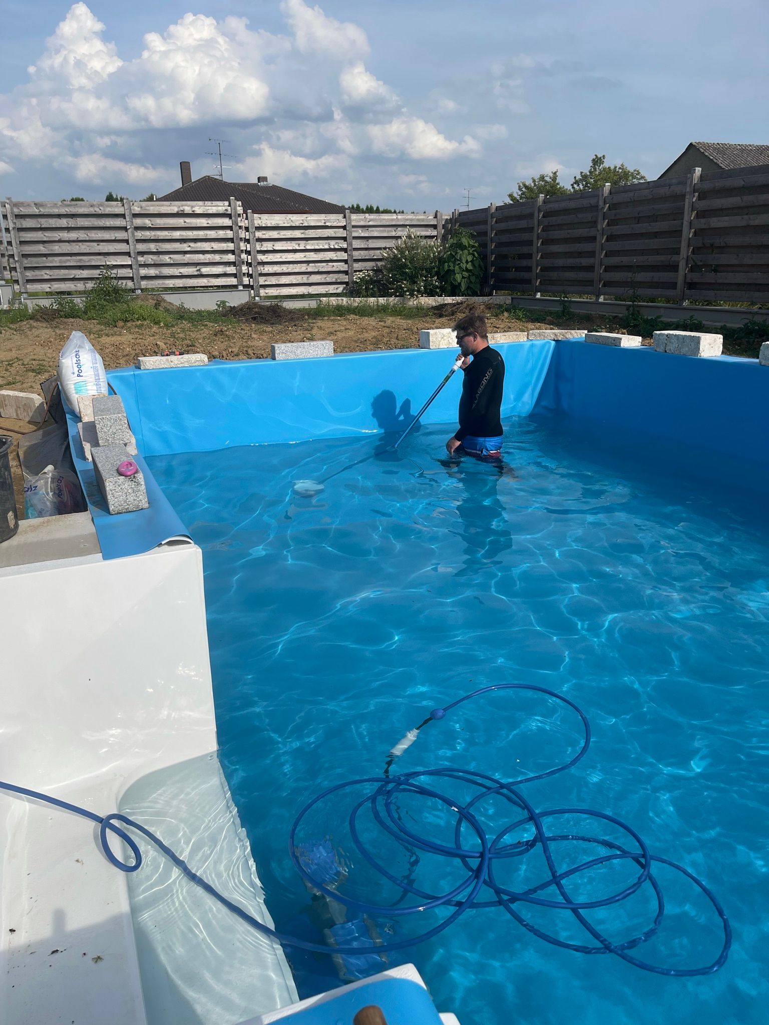 Ein Kind reinigt einen Pool mit einem Schlauch. Blaues Wasser und Himmel, weiße Beckenränder. Im Hintergrund ein Zaun.