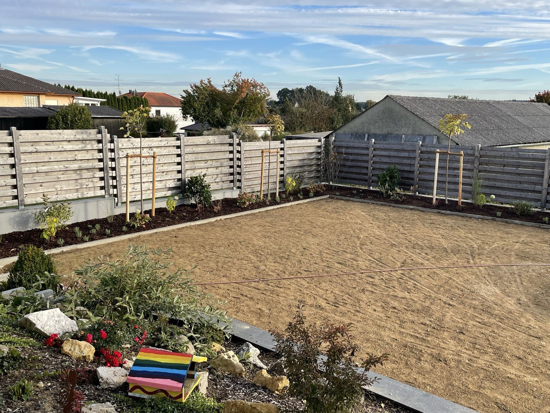 Ein Hinterhof mit einem Holzzaun und frisch ausgelegtem Mulch. Im Hintergrund sind Bäume und Häuser unter einem blauen Himmel zu sehen.