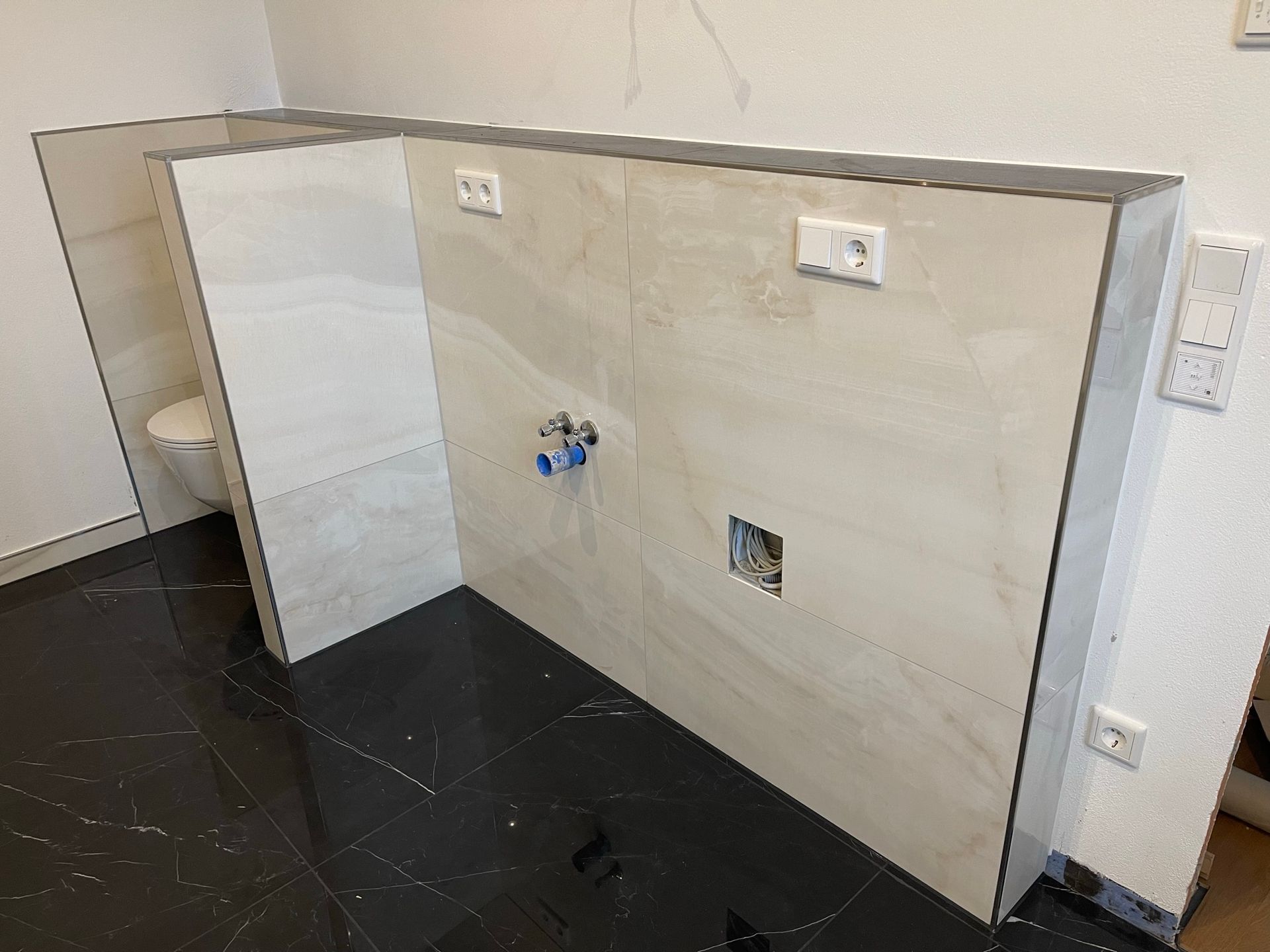 Badezimmerwand mit hellbeigen Fliesen, schwarzer Boden, Toilette teilweise sichtbar. Steckdosen und Sanitärarmaturen.