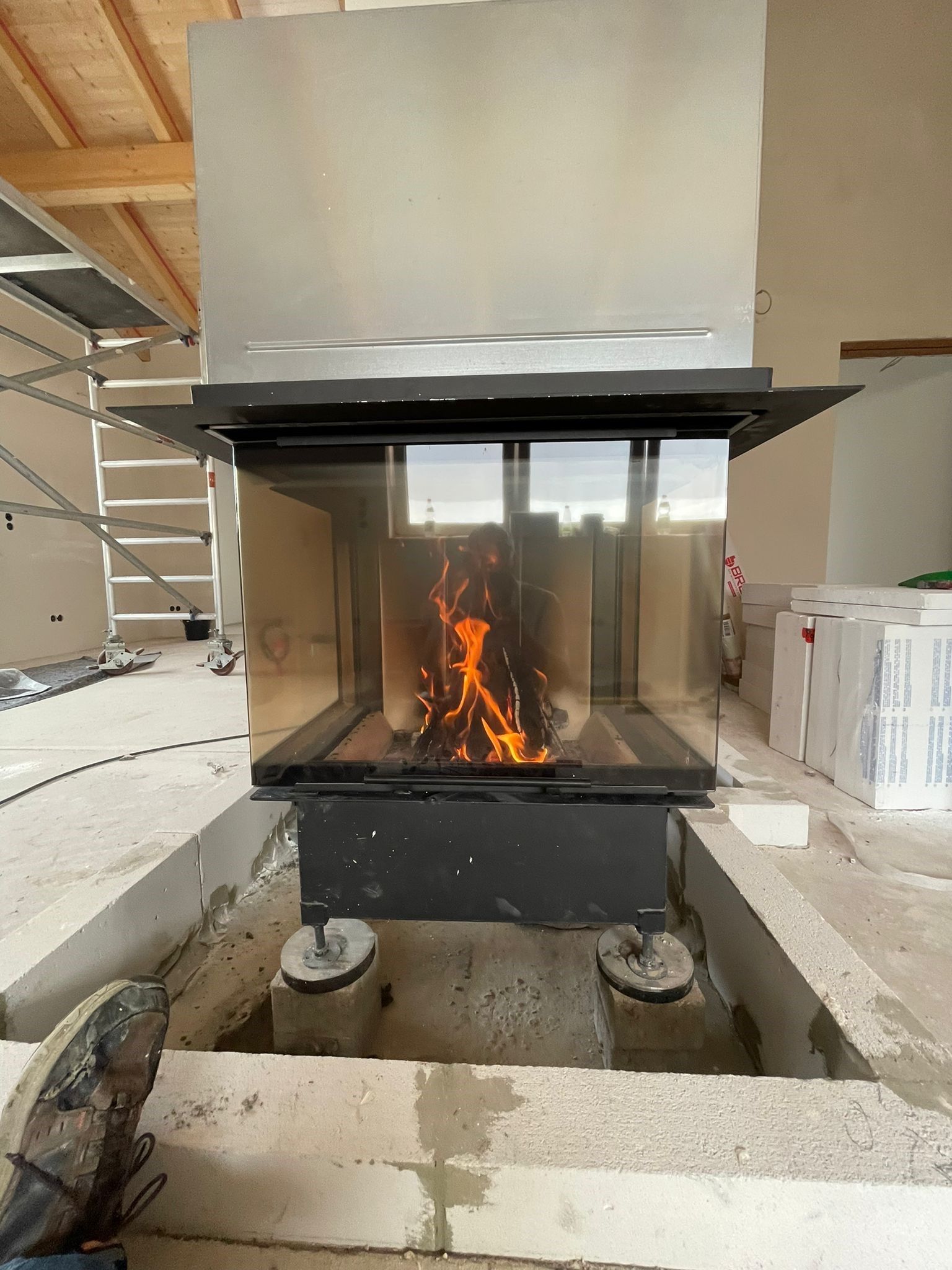 Kamin im Bau, Flammen sichtbar. Schwarzer Rahmen, Glasplatten. Eingebaut in eine Betonkonstruktion, unfertiger Raum.