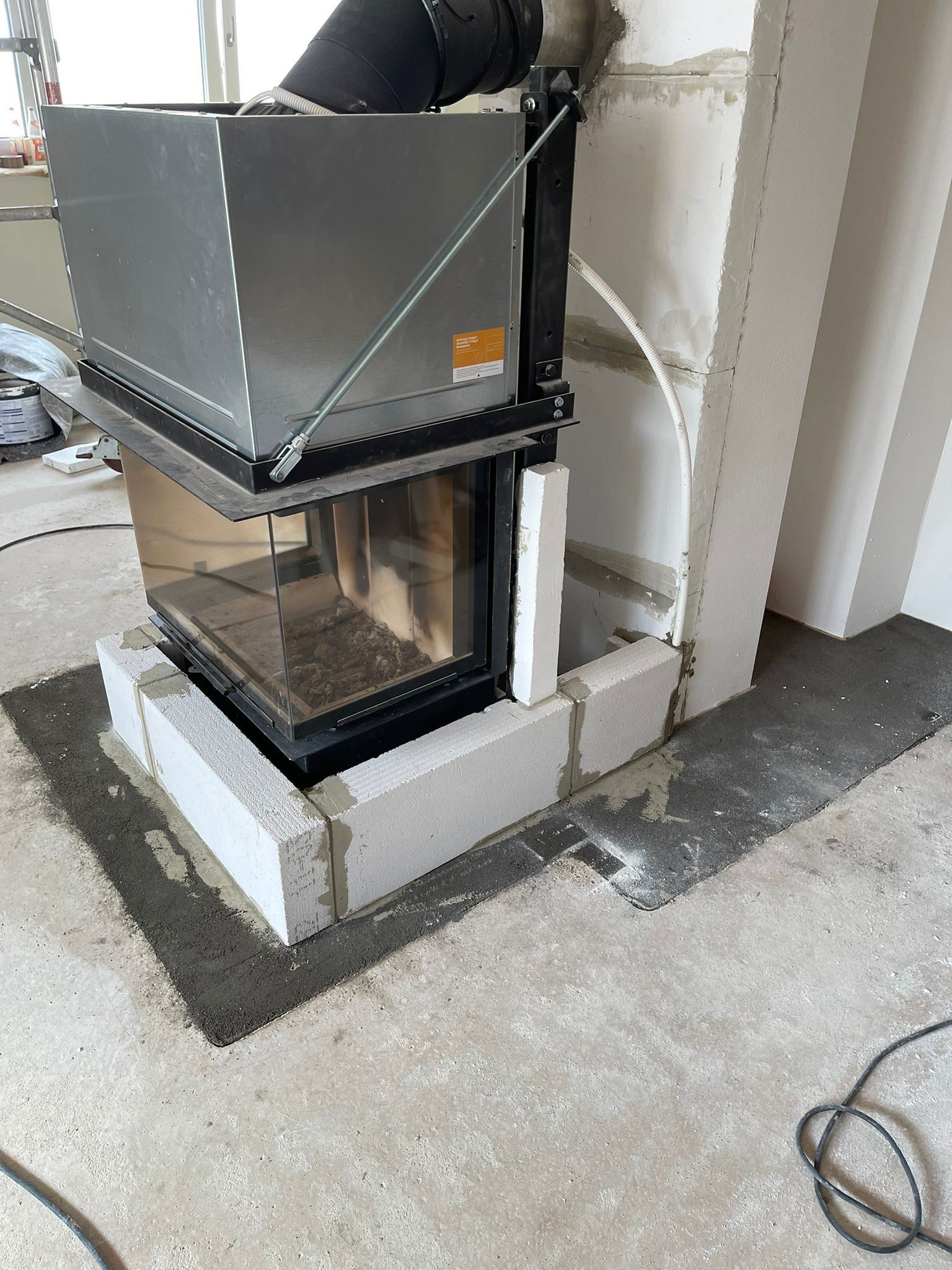 Kamininstallation: Ein grauer Metallkamin wird in einem Raum in weißen Schlackenblöcken auf einem Betonsockel eingelassen.