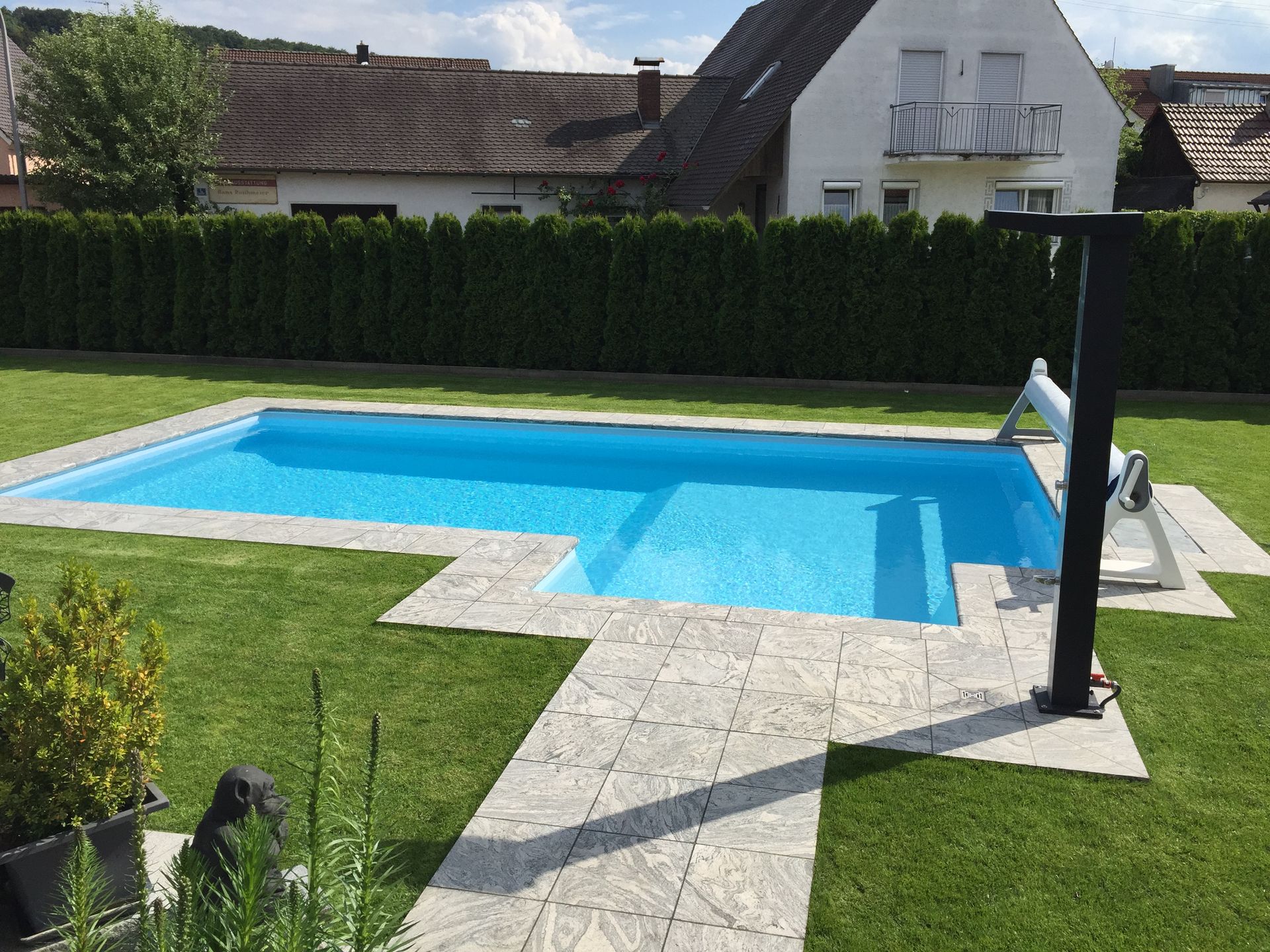 Pool im Hinterhof, blaues Wasser, Steinterrasse, grünes Gras, ein Haus und eine Hecke.