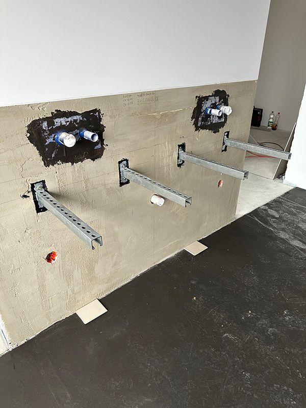 An einer Wand mit Rohrleitungen montierte Halterungen, wahrscheinlich für einen Badezimmerwaschtisch. Braune Wand und dunkler Boden, Baustelle.