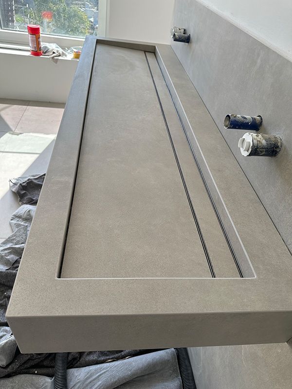 Grauer Waschtisch aus Beton mit rechteckigem Waschbecken, an der Wand montiert. Zwei Wasserhähne sichtbar.