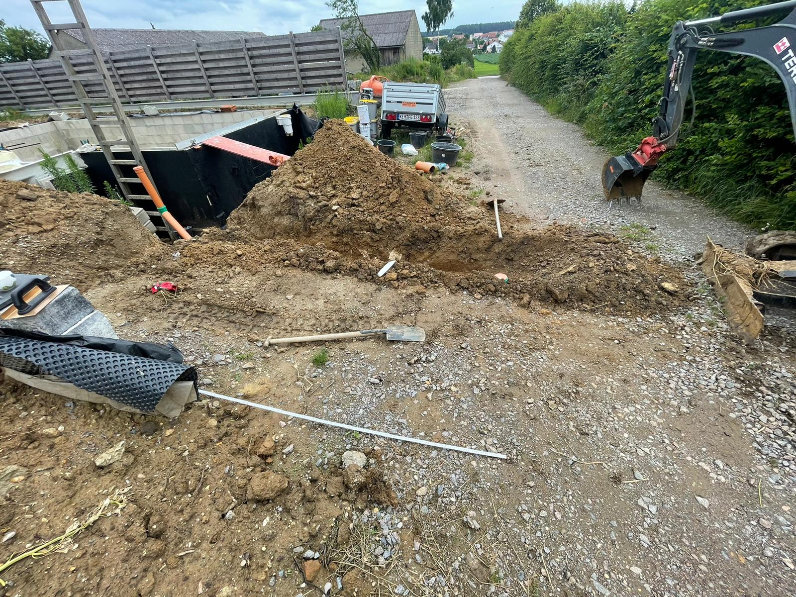 Baustelle mit einem Bagger, der in einem Feldweg gräbt; Materialien und ein Anhänger sind vorhanden.