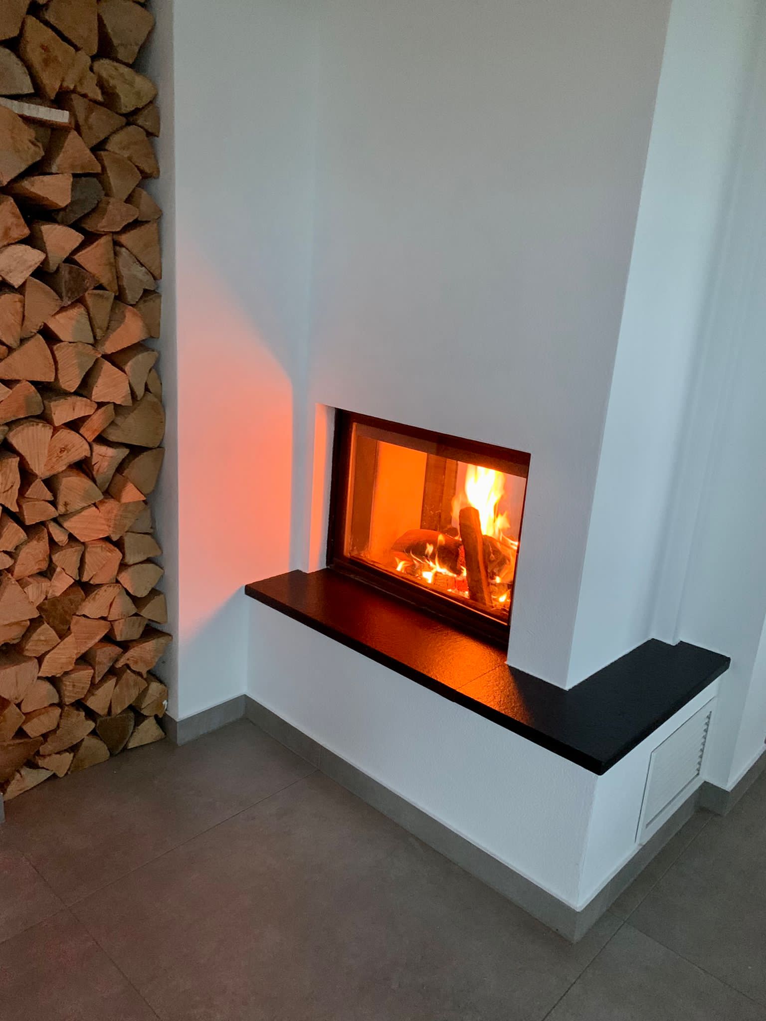 Kamin mit brennendem Feuer, weißen Wänden, schwarzem Herd und Brennholzstapel.