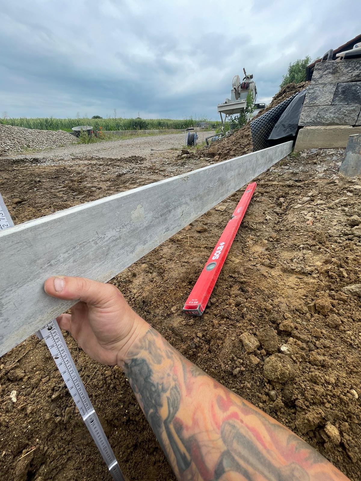 Person, die auf einer Baustelle eine Betonschalung hält; außerdem sind eine Wasserwaage und ein Maßband vorhanden.