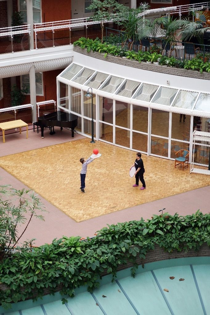 Kaksi ihmistä heittelee palloa sisäpihalla, jossa on lasikatto, kasveja ja istuimia ; Activus Oy:n terapiaympäristö