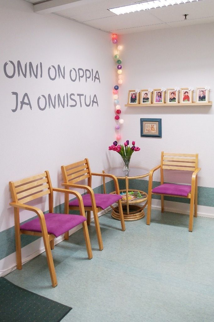 Odotushuone: kolme puista tuolia violeteilla tyynyillä, maljakko kukkia, hylly kehystettyine valokuvineen ja seinällä teksti Onni on oppia ja onnistua. 