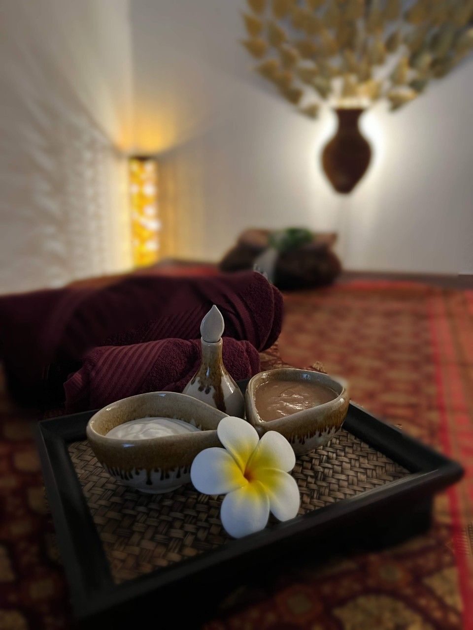 Ein Spa-Tablett mit Schälchen mit Kräuterpaste und einer weissen Blüte auf einer gemusterten Matte in einem sanft beleuchteten Raum