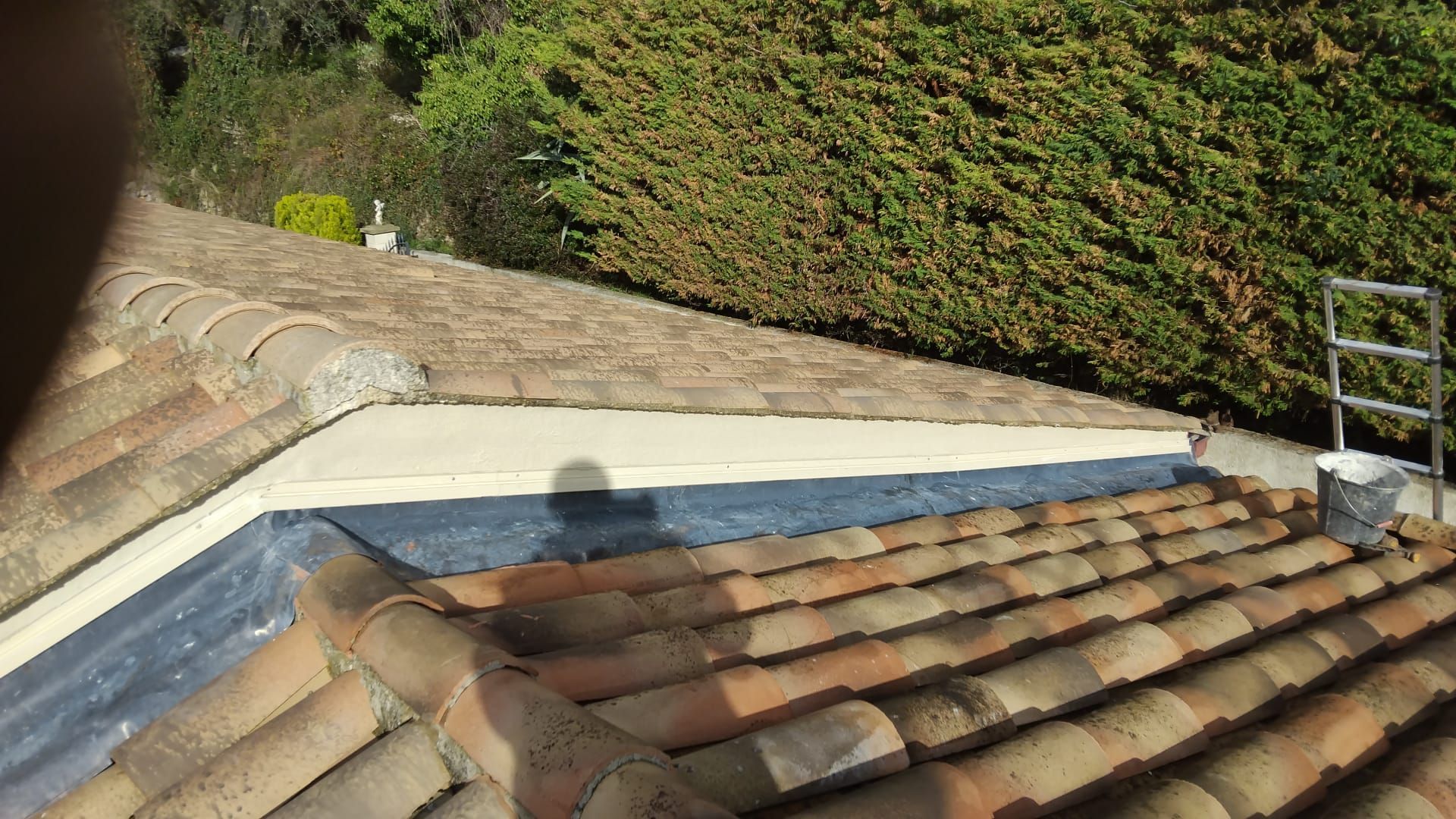 Toiture en tuiles de terre cuite, parapet blanc et colline verdoyante en arrière-plan.