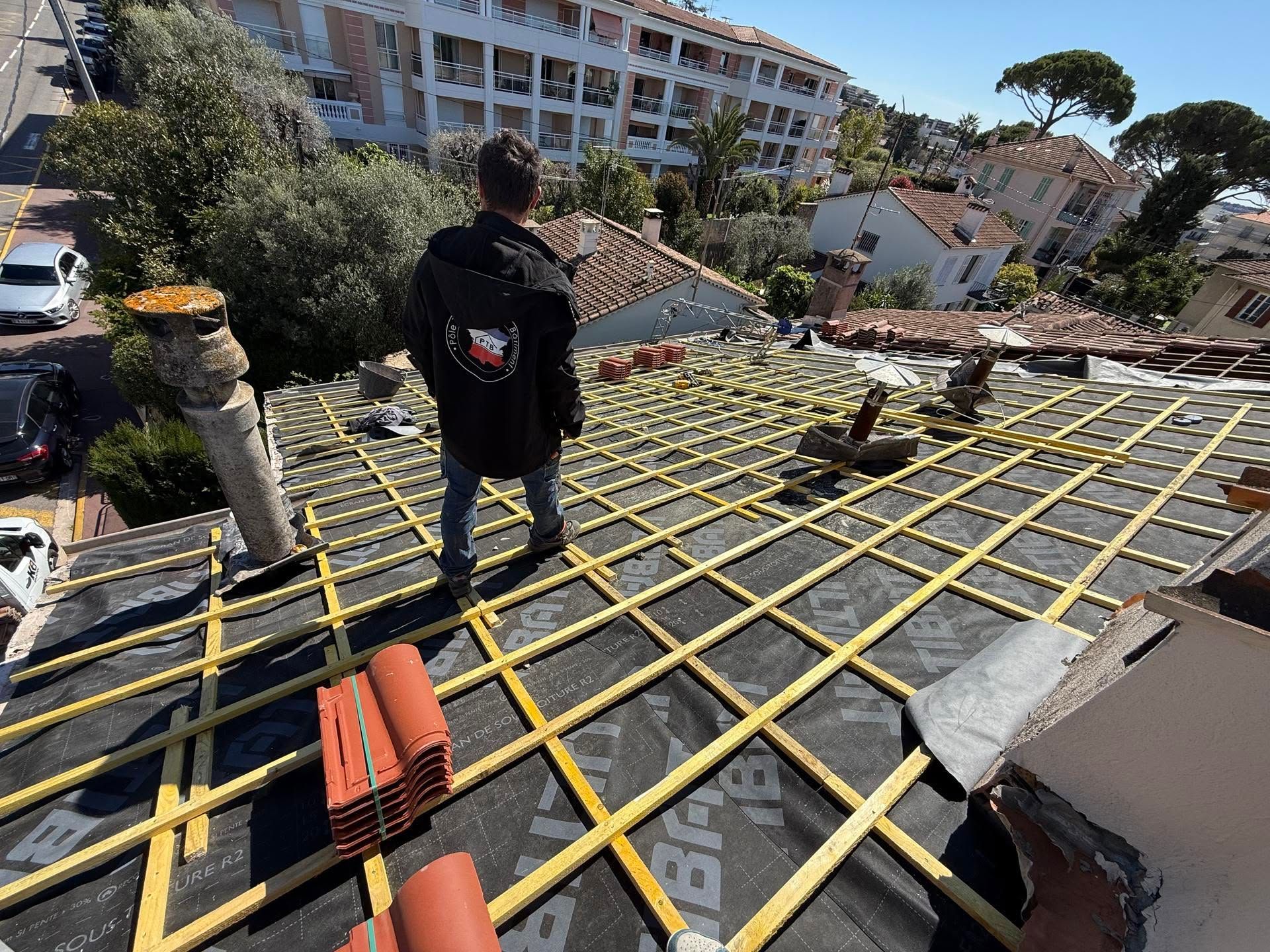 Un ouvrier se tient sur un toit en construction, avec une charpente et des matériaux de construction à proximité.