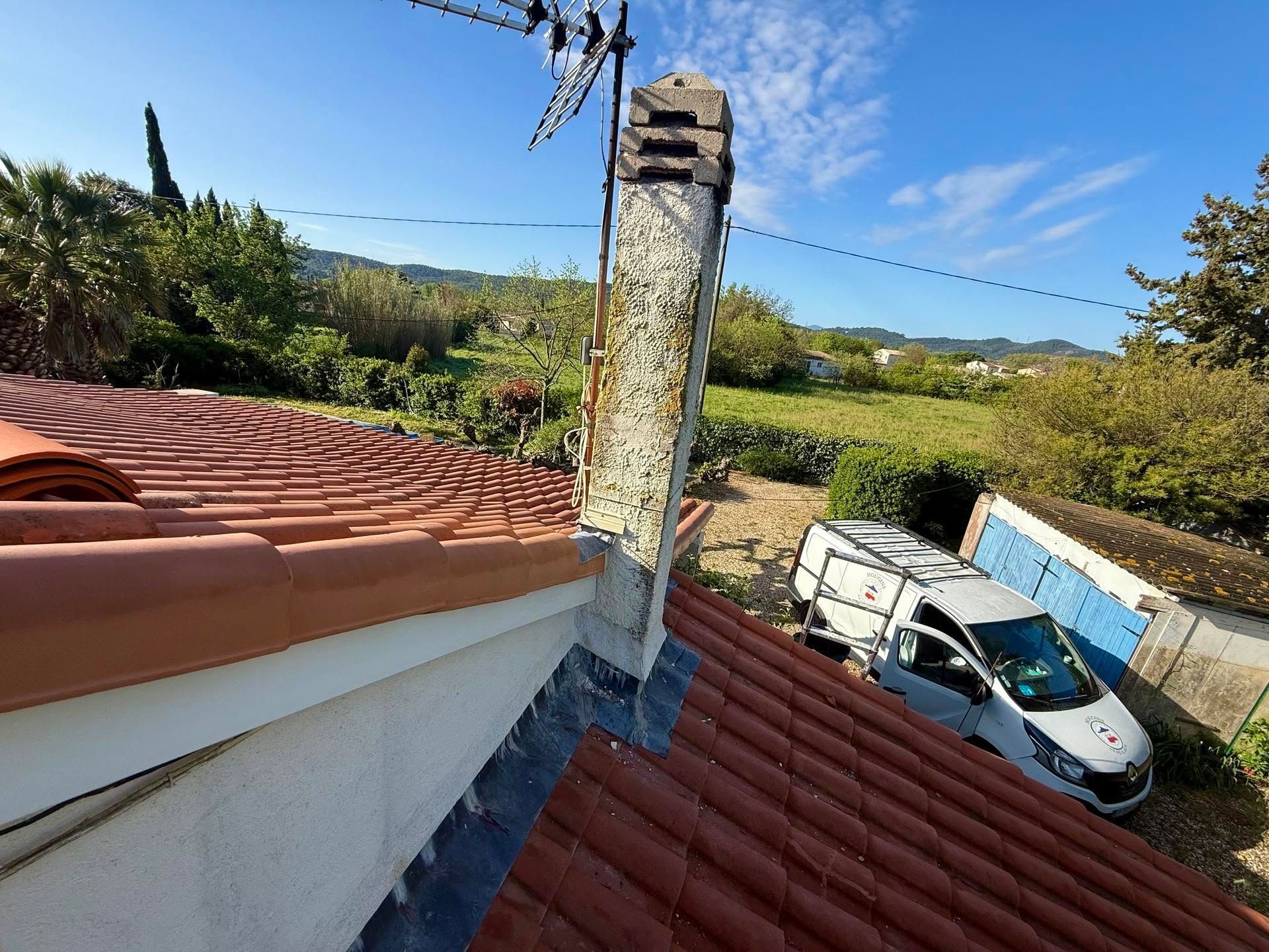 Vue depuis le toit avec des tuiles rouges, une cheminée patinée et une voiture blanche garée en contrebas, sous un ciel bleu.