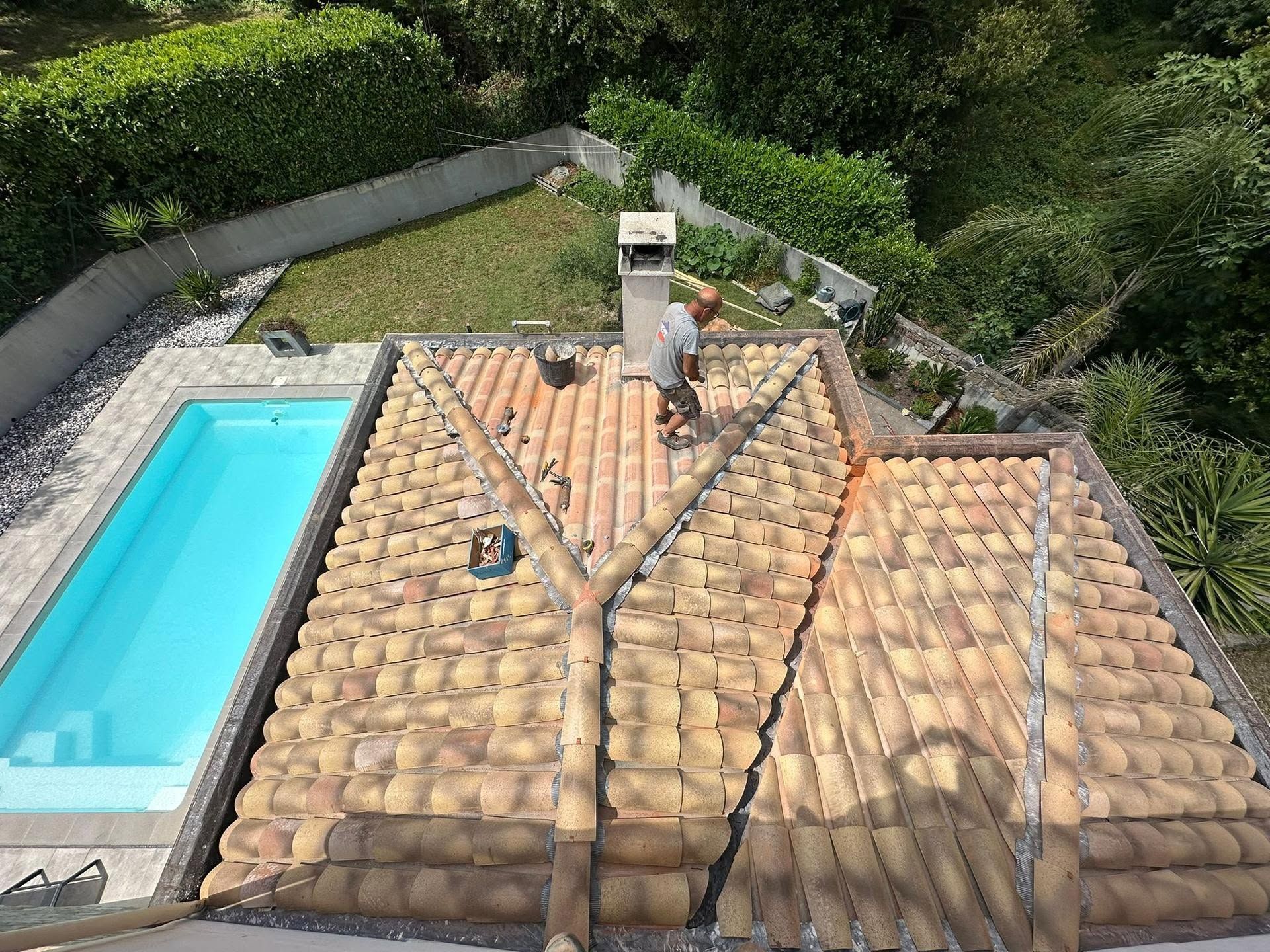 Des couvreurs travaillent sur un toit de tuiles à côté d'une petite piscine et d'un aménagement paysager verdoyant.