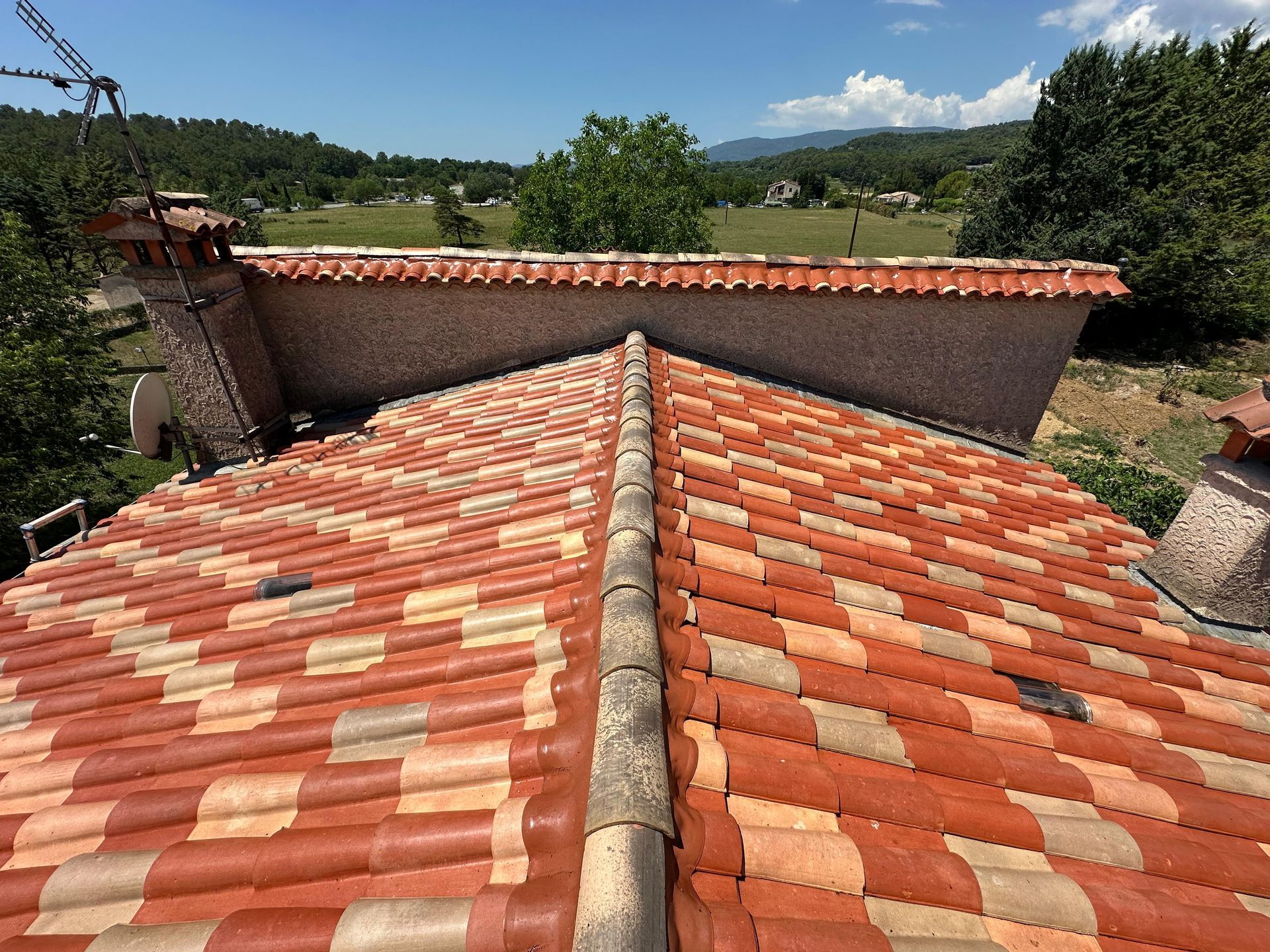 Au premier plan, un toit de tuiles en terre cuite rouge, un bâtiment en pierre et une campagne verdoyante sous un ciel bleu.