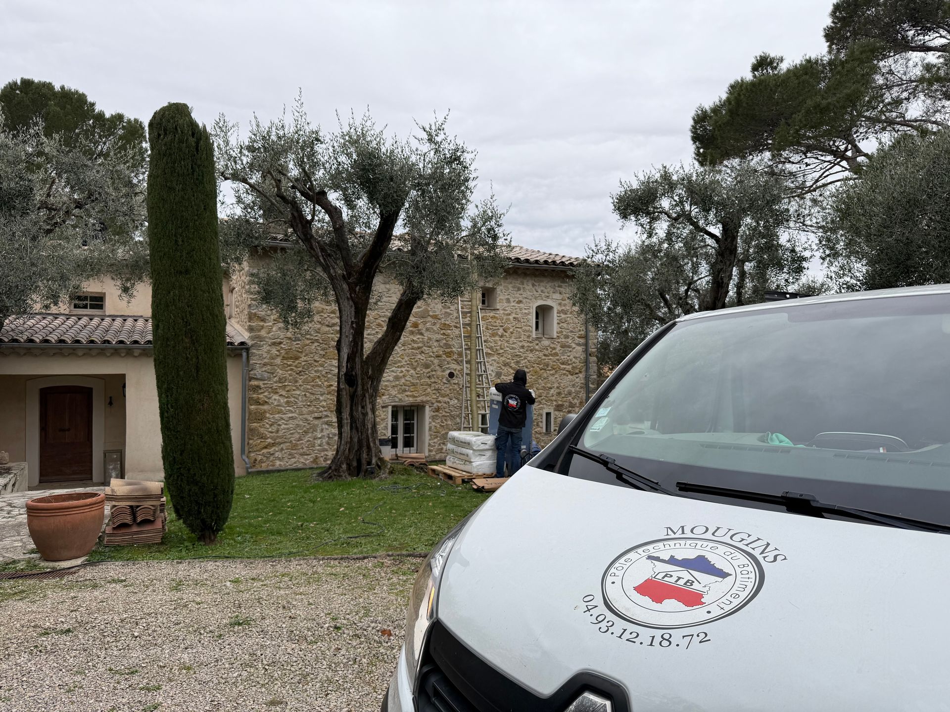 Une camionnette blanche avec un logo au premier plan, à côté d'une maison en pierre et d'un jardin sous un ciel couvert.