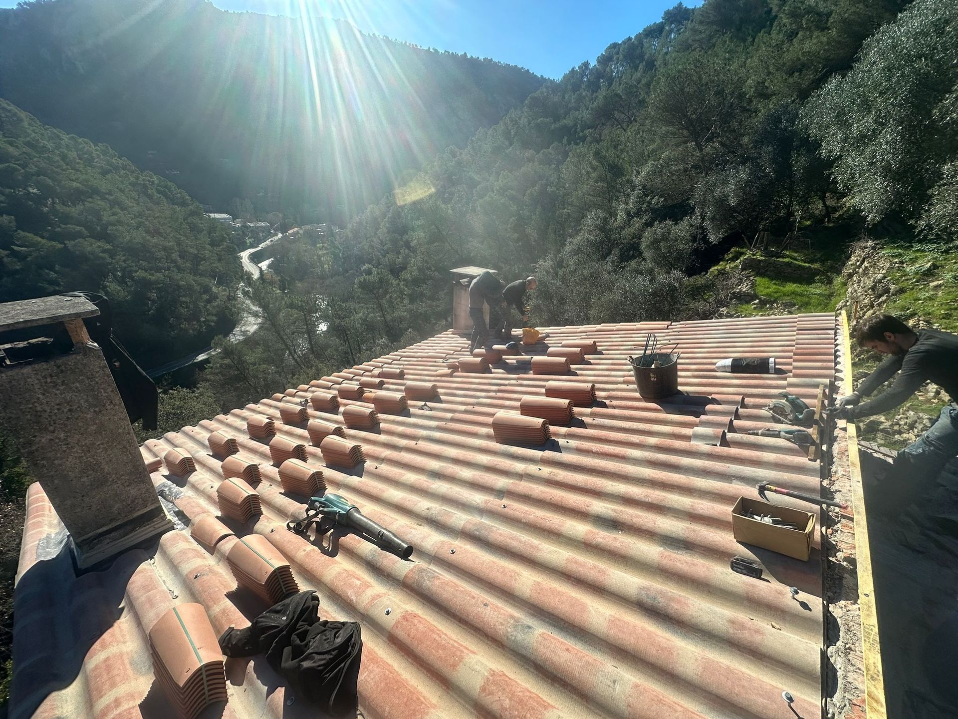 Des ouvriers travaillent sur un toit en tôle ondulée à flanc de colline boisée, sous un soleil éclatant.