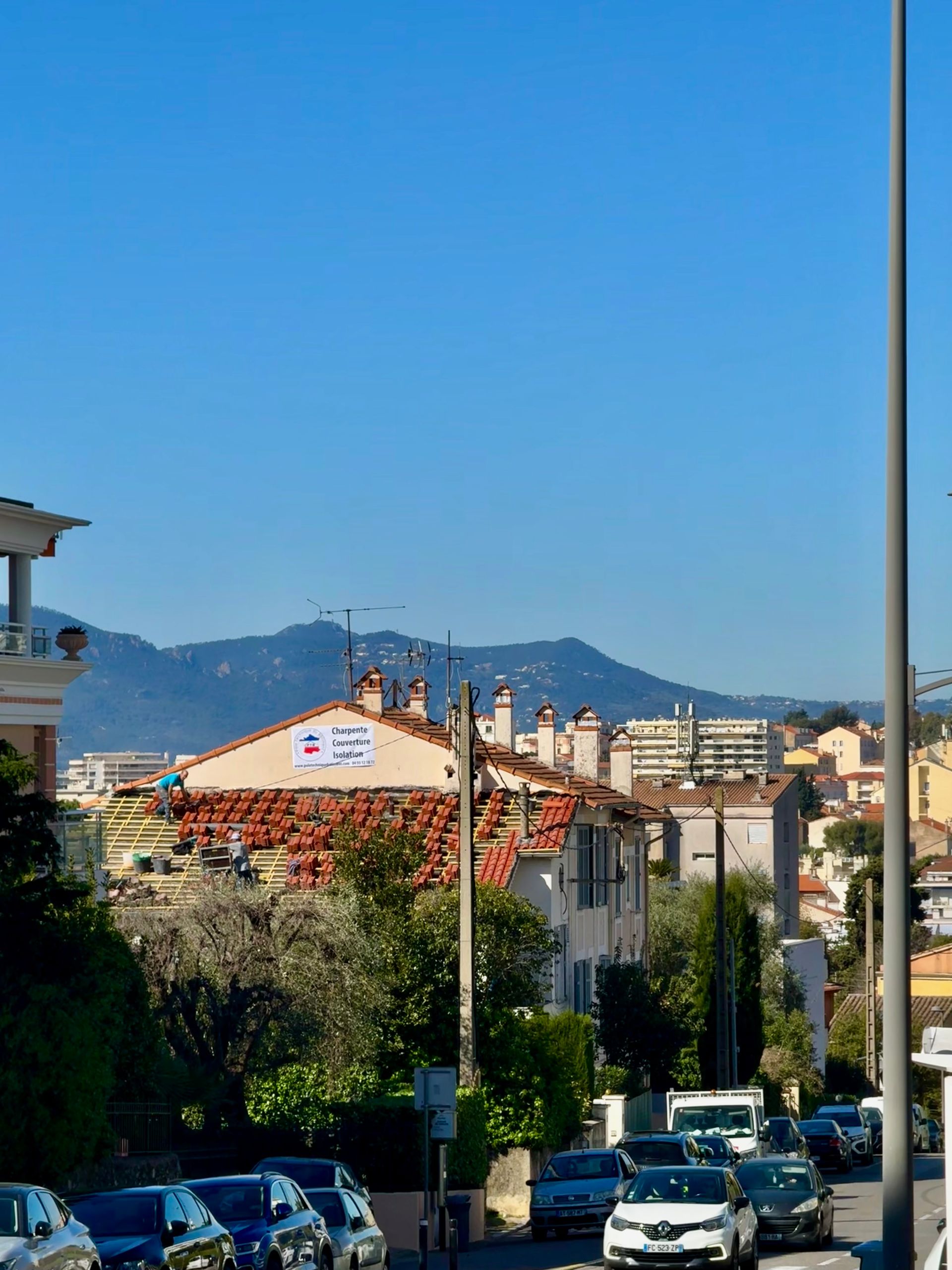 Rue urbaine avec voitures garées, bâtiments aux toits rouges et montagnes sous un ciel bleu limpide.