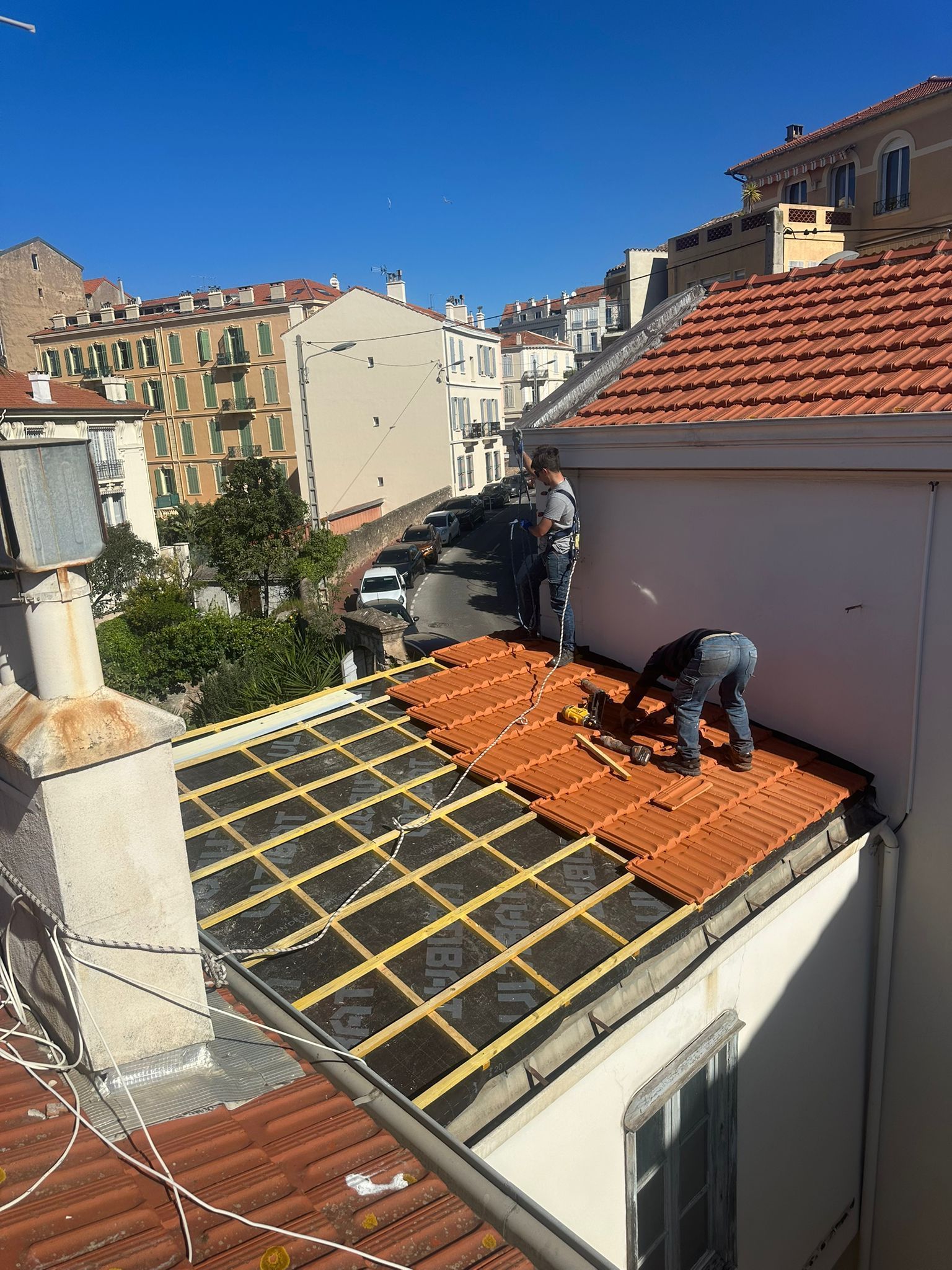 Un ouvrier pose des tuiles sur un toit plat, à côté d'un toit de tuiles rouges, en milieu urbain.