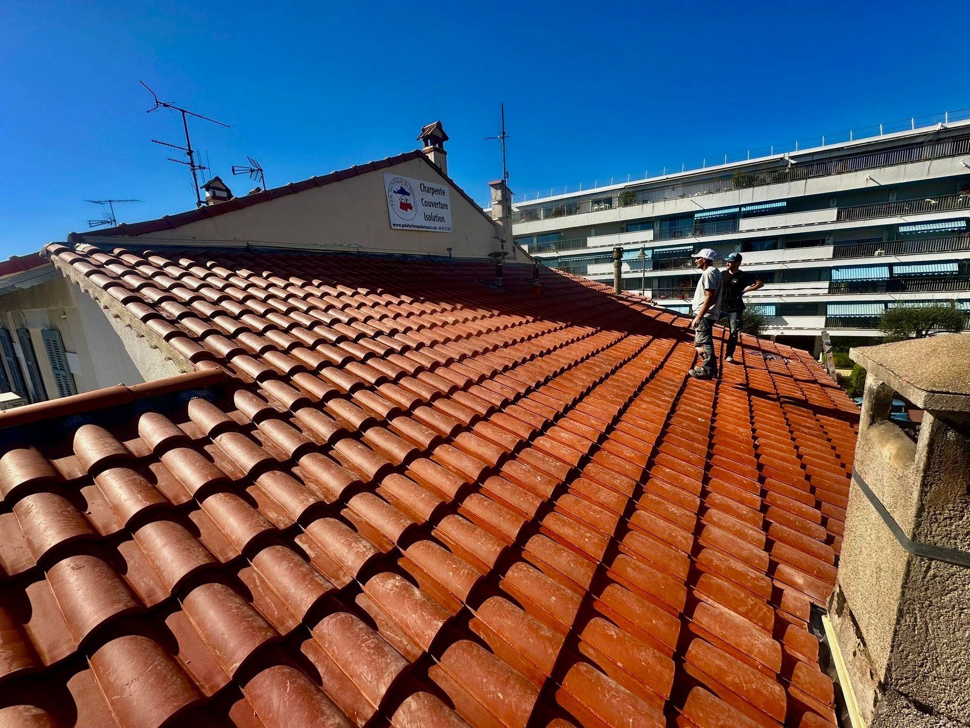 Deux ouvriers travaillent sur un toit de tuiles rouges.