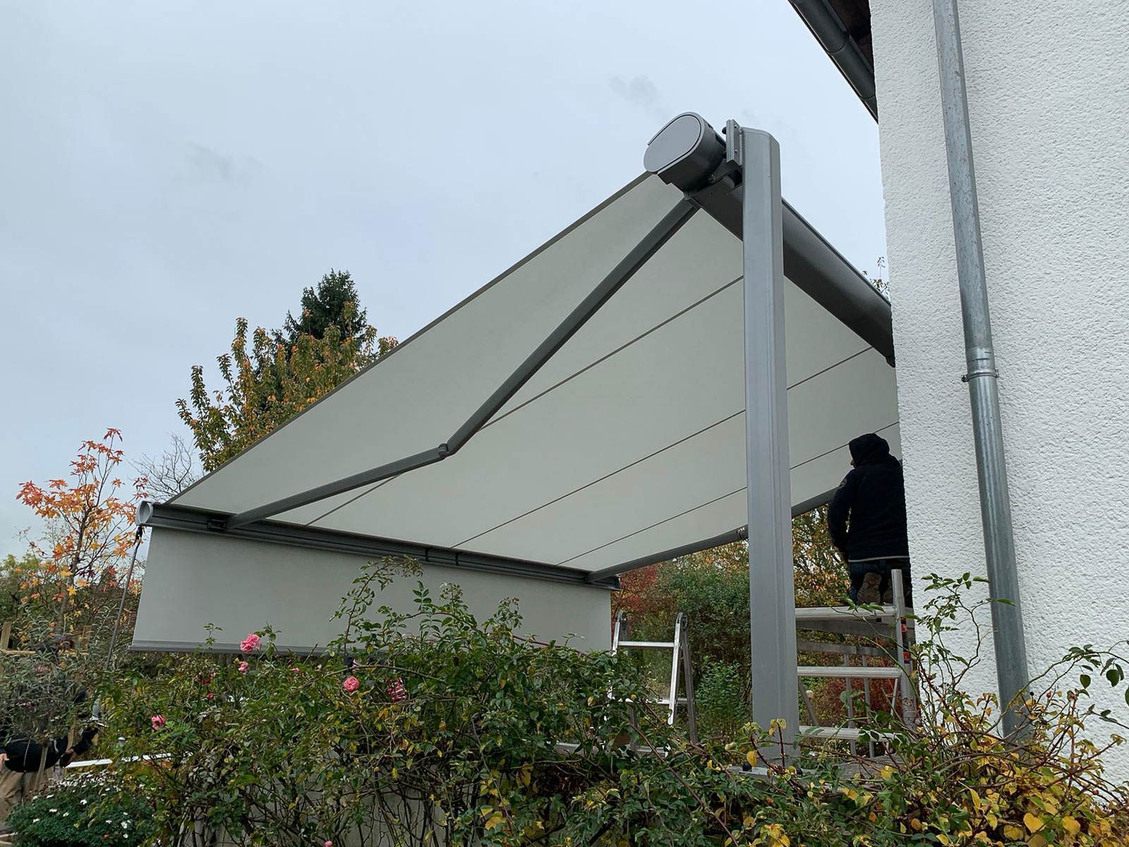 Eine Person steht auf einer Leiter unter einem Vordach an der Seite eines Gebäudes.