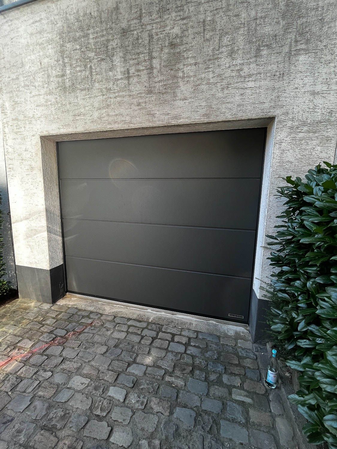 An der Seite eines Gebäudes neben einer gepflasterten Auffahrt befindet sich ein schwarzes Garagentor.